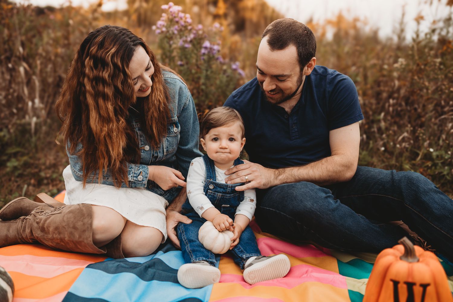 Young family sitting on colorful blanket in autumn field with baby and small pumpkin.