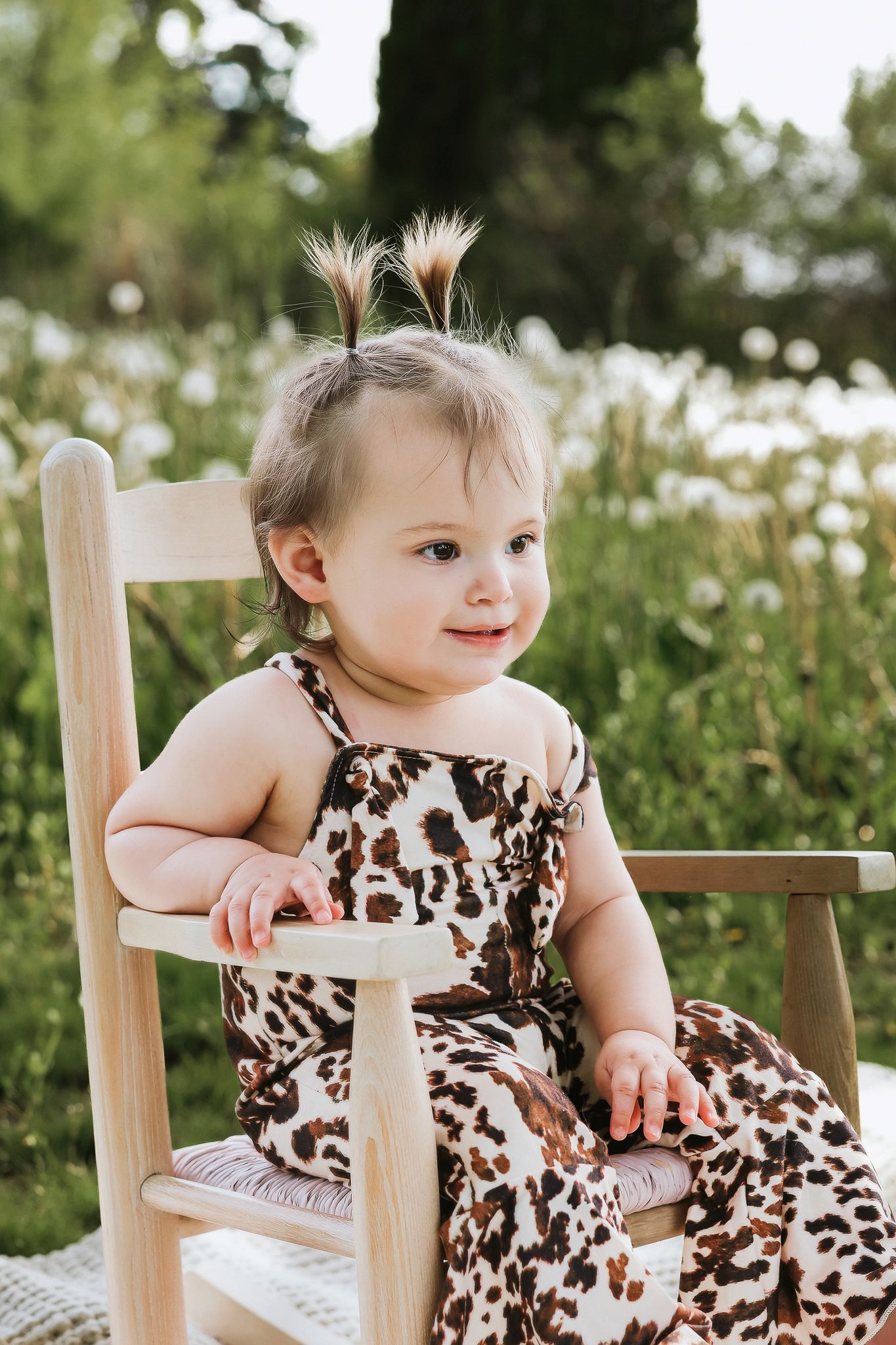 Adorable baby girl in leopard print outfit sitting in a wooden chair outdoors in a flower field.