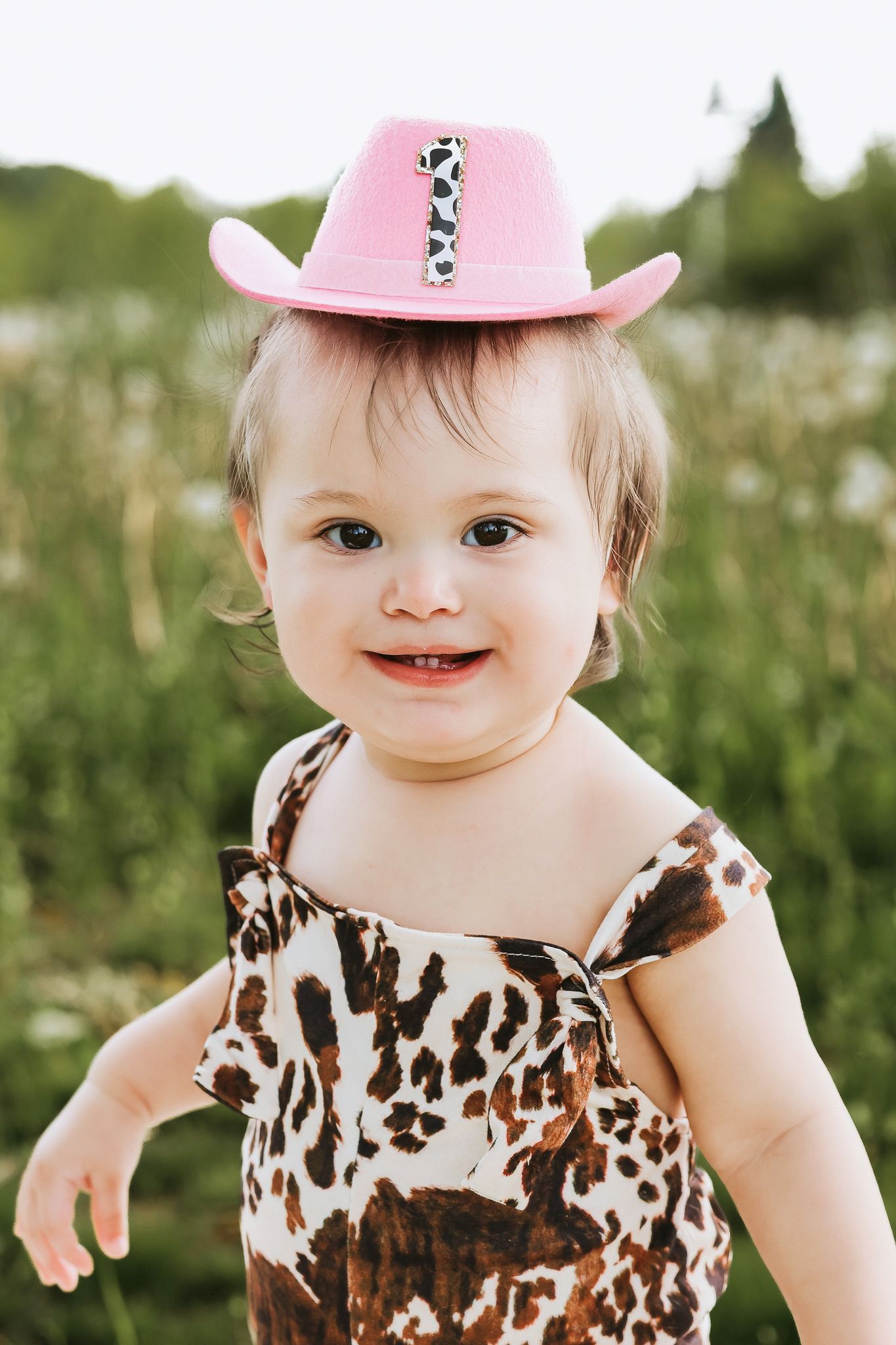 Cute baby girl wearing pink cowboy hat and animal print romper smiling outdoors.