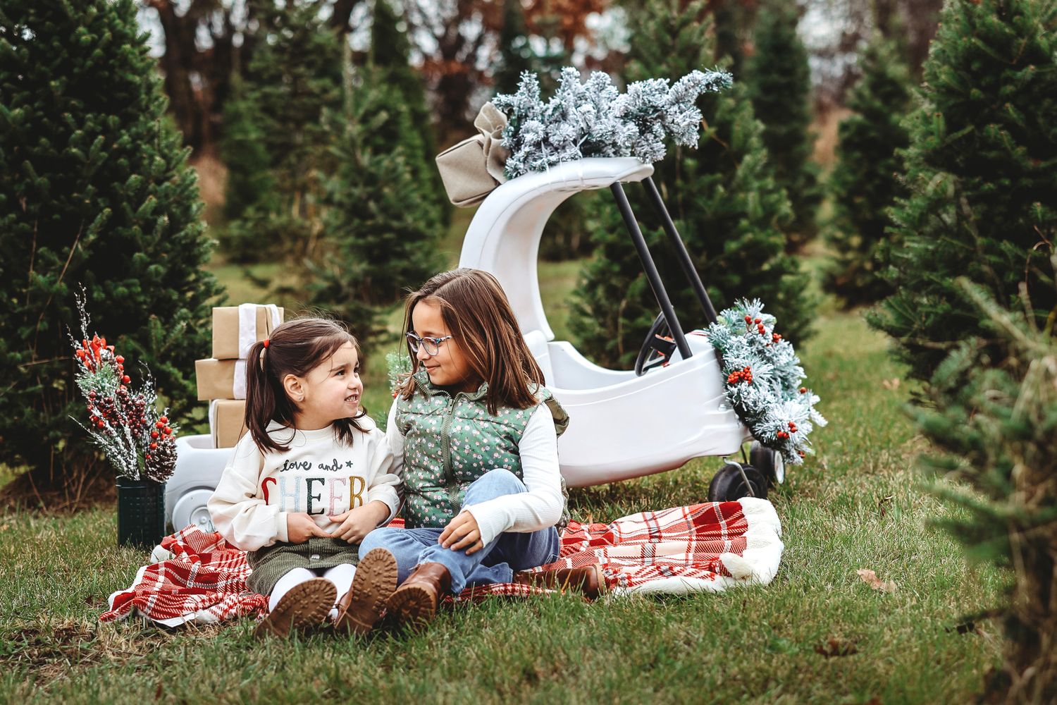 Two young sisters sit together on a plaid blanket near a festive sleigh at a Christmas tree farm.