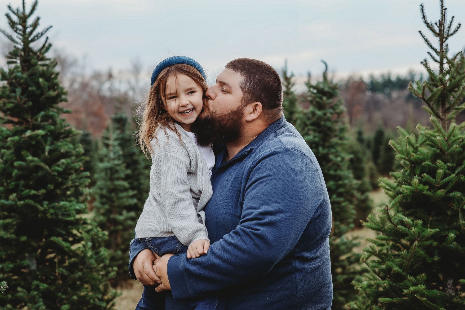 Bearded father kisses smiling little girl wearing blue beret at a Christmas tree farm.