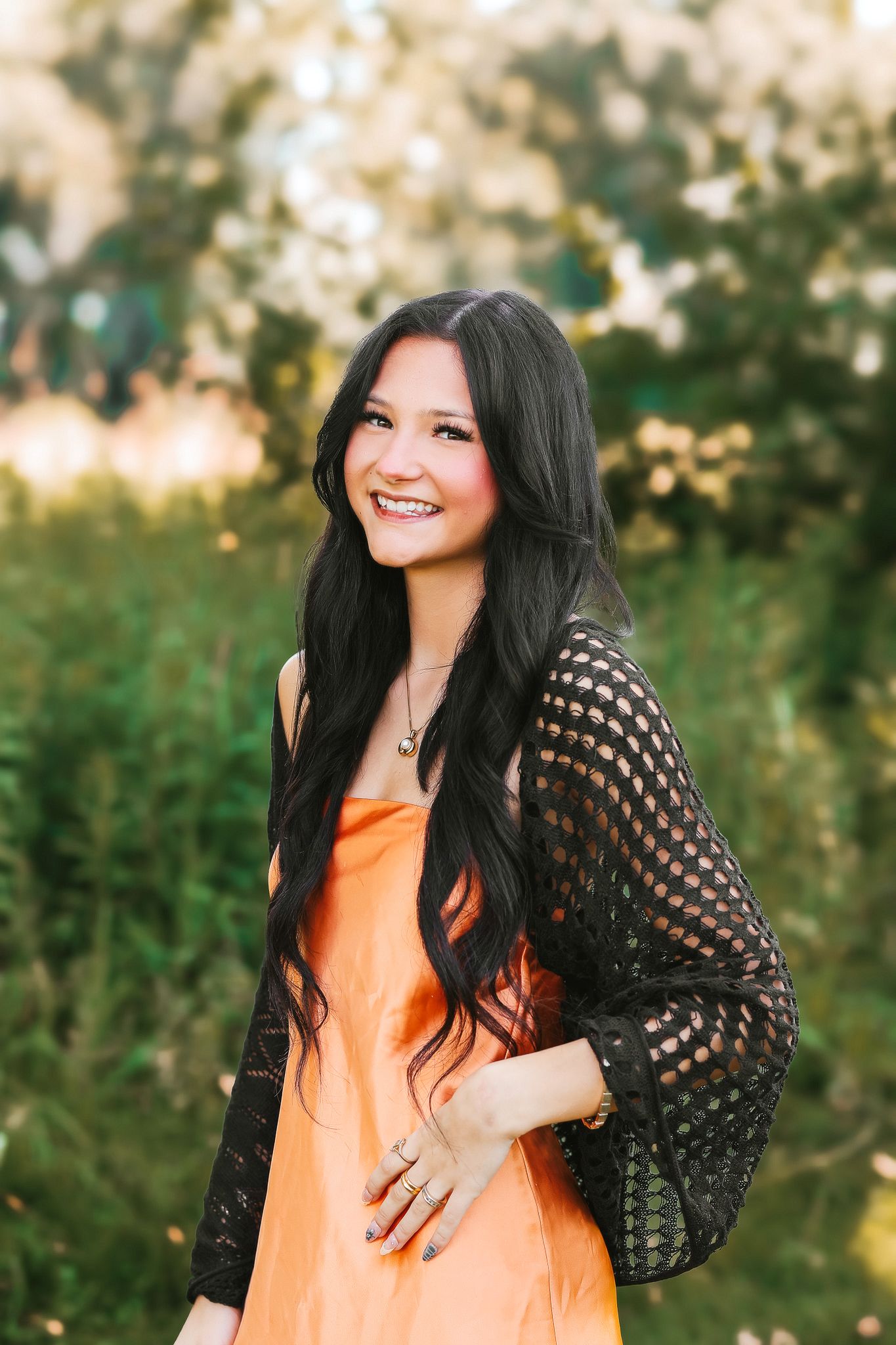 Smiling teenage girl with long dark hair in an orange dress and black lace cardigan posing in a sunny garden.