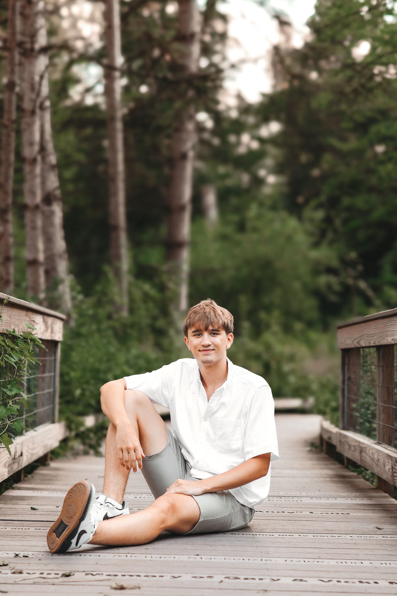 Young man in white shirt and gray shorts sitting relaxed on a wooden bridge in a green forest setting.