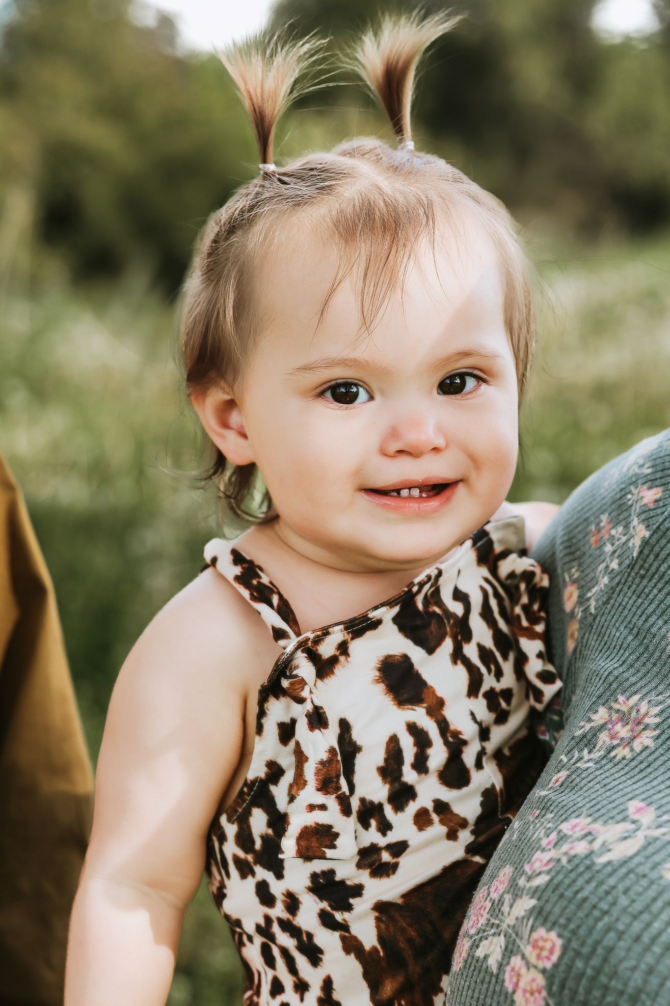 Cheerful baby girl with tiny pigtails in leopard print outfit being held outdoors in a green field.