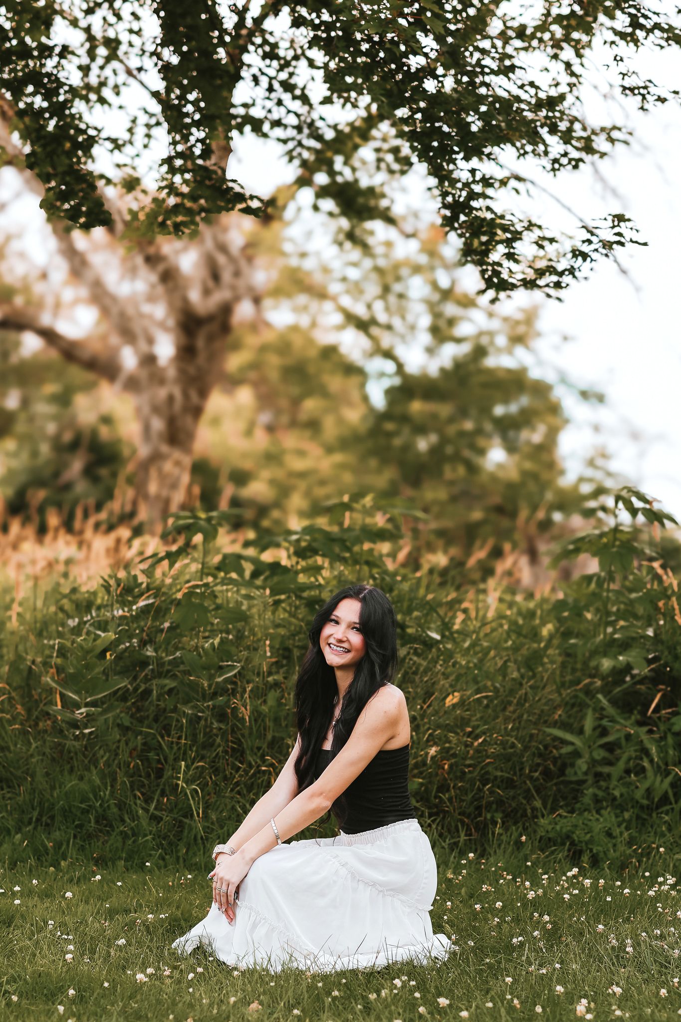 Smiling young woman with dark hair sitting on grass in a white skirt and black top beneath a large tree outdoors.