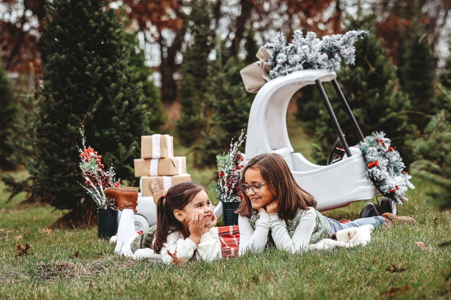 Two girls lying on grass surrounded by Christmas gifts and holiday decorations outdoors.