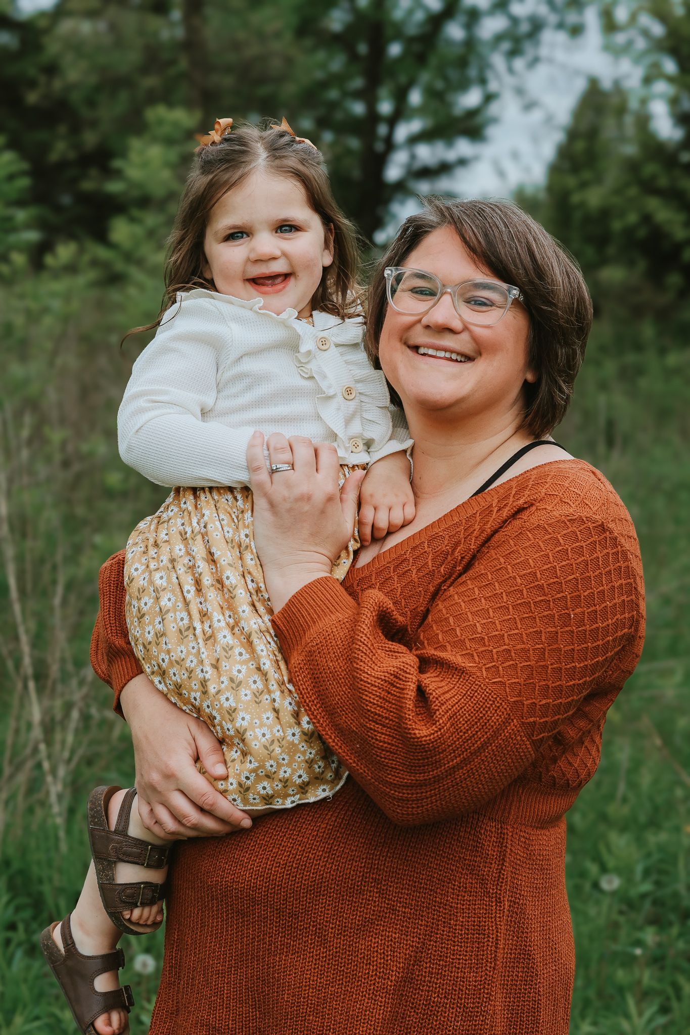 A smiling woman in an orange sweater holds a happy young girl outdoors in a green field.