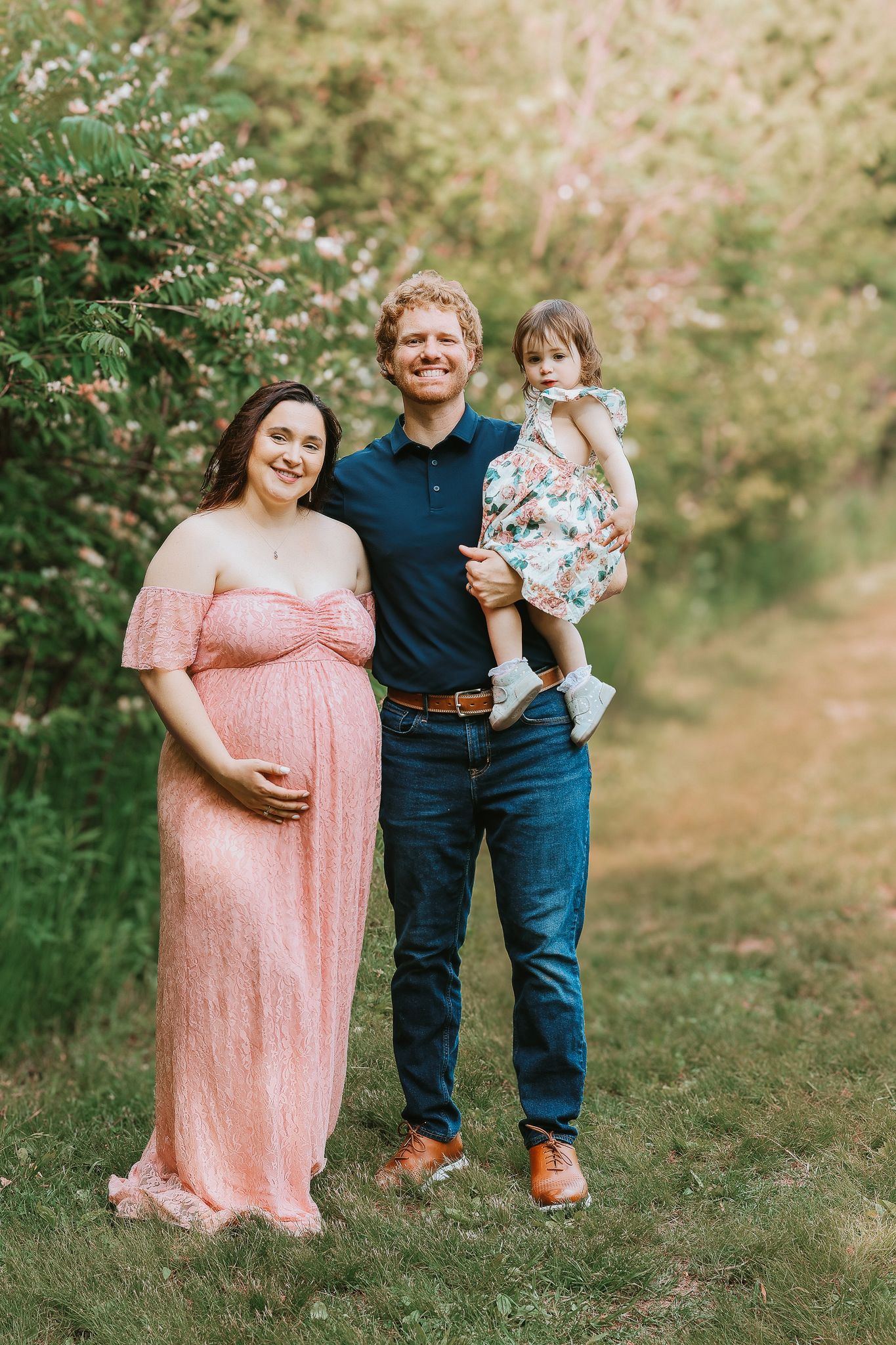 Pregnant woman in pink lace gown with husband and toddler daughter in spring garden.
