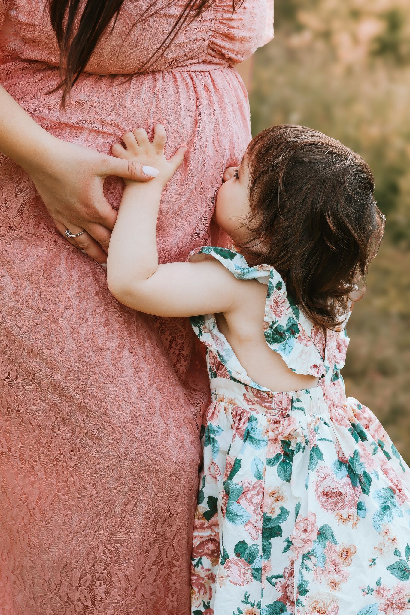 Toddler girl in floral dress kissing pregnant mothers baby bump outdoors.