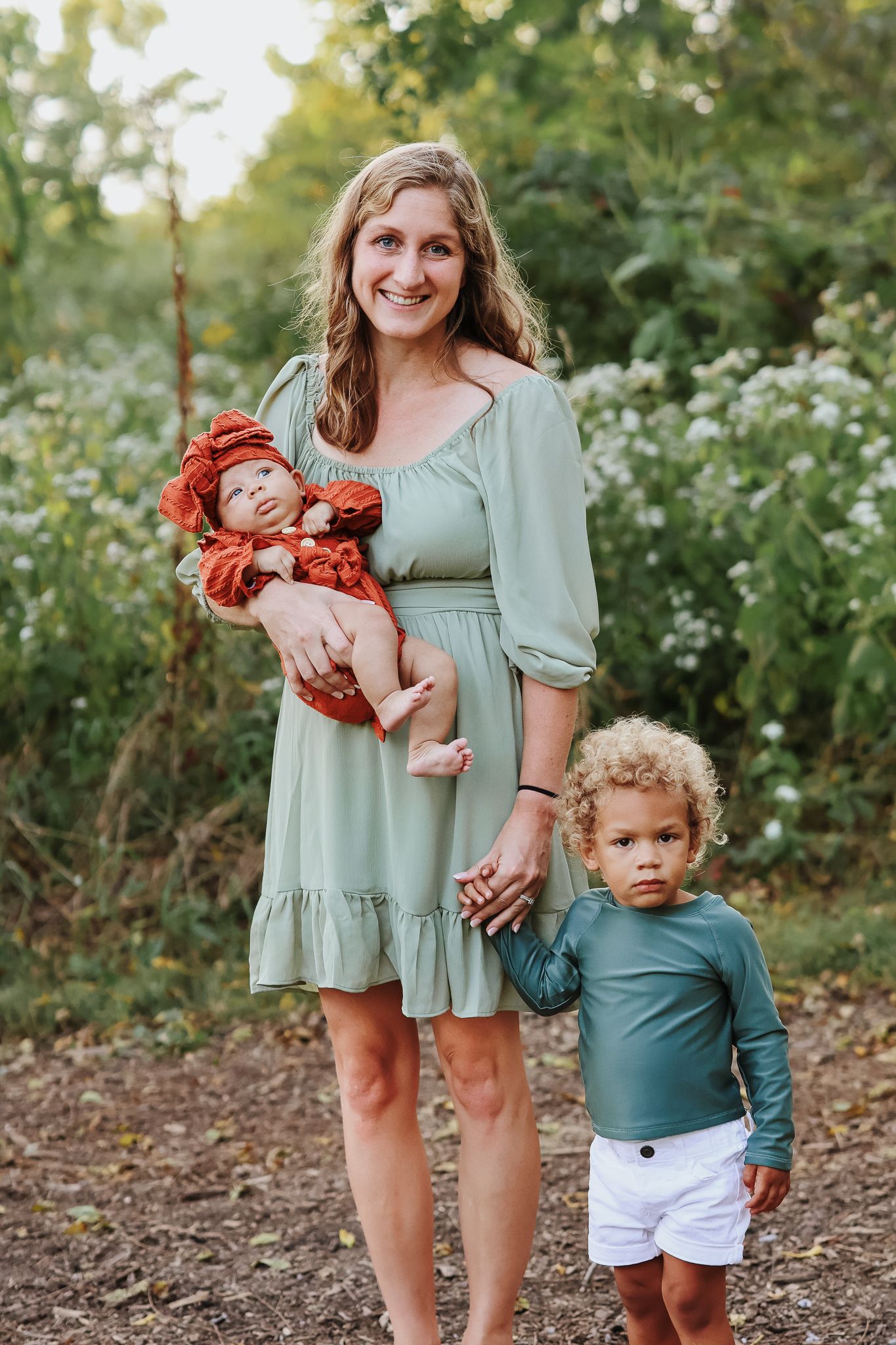 Smiling mother in a sage green dress holding a newborn and standing with her curly-haired toddler outdoors.