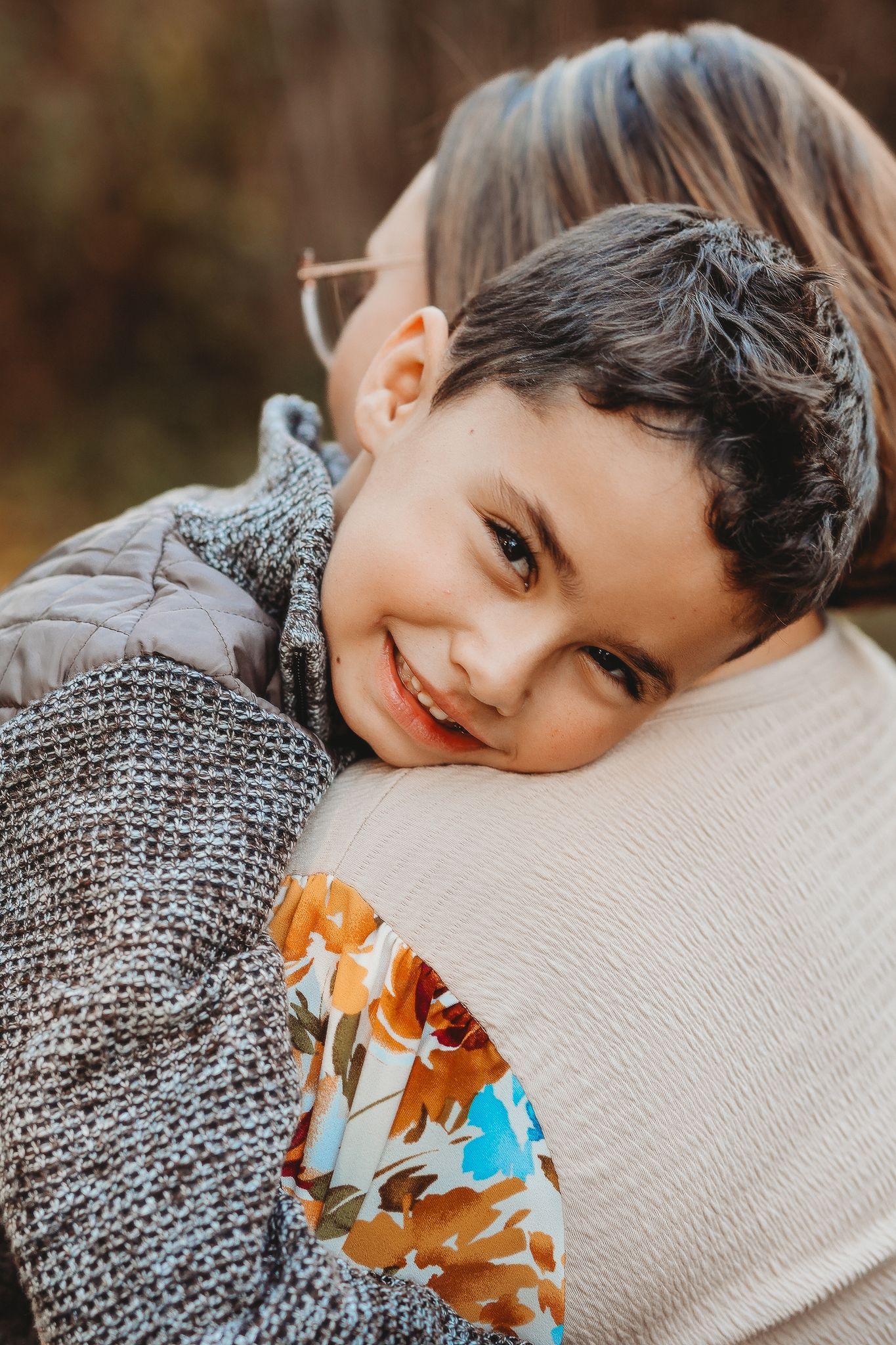 A smiling young boy rests his head on his mothers shoulder in a warm embrace outdoors.