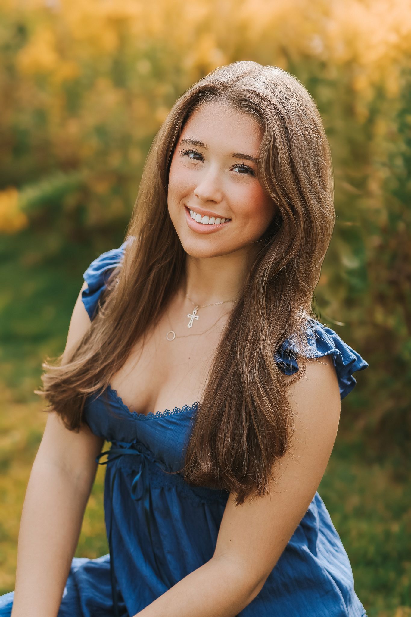 Close-up portrait of smiling brunette teen girl wearing blue dress and cross necklace outdoors.
