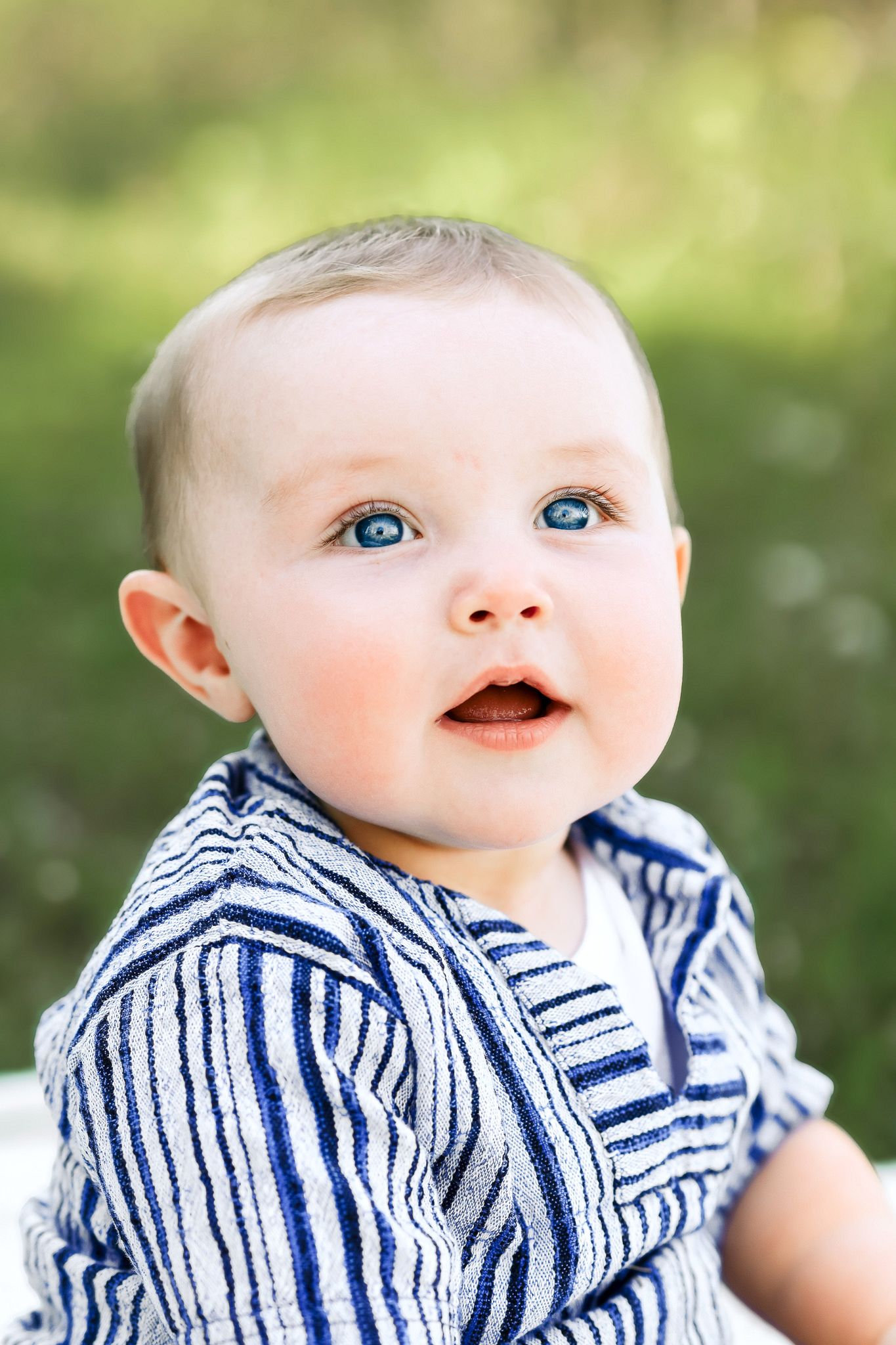 Happy blue-eyed baby boy in striped navy jacket looking up curiously outdoors in green park.