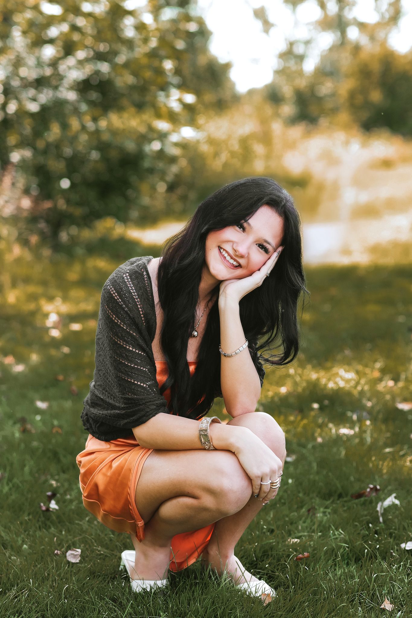Smiling young woman with long dark hair crouching on grass in an orange skirt and dark knit cardigan outdoors.