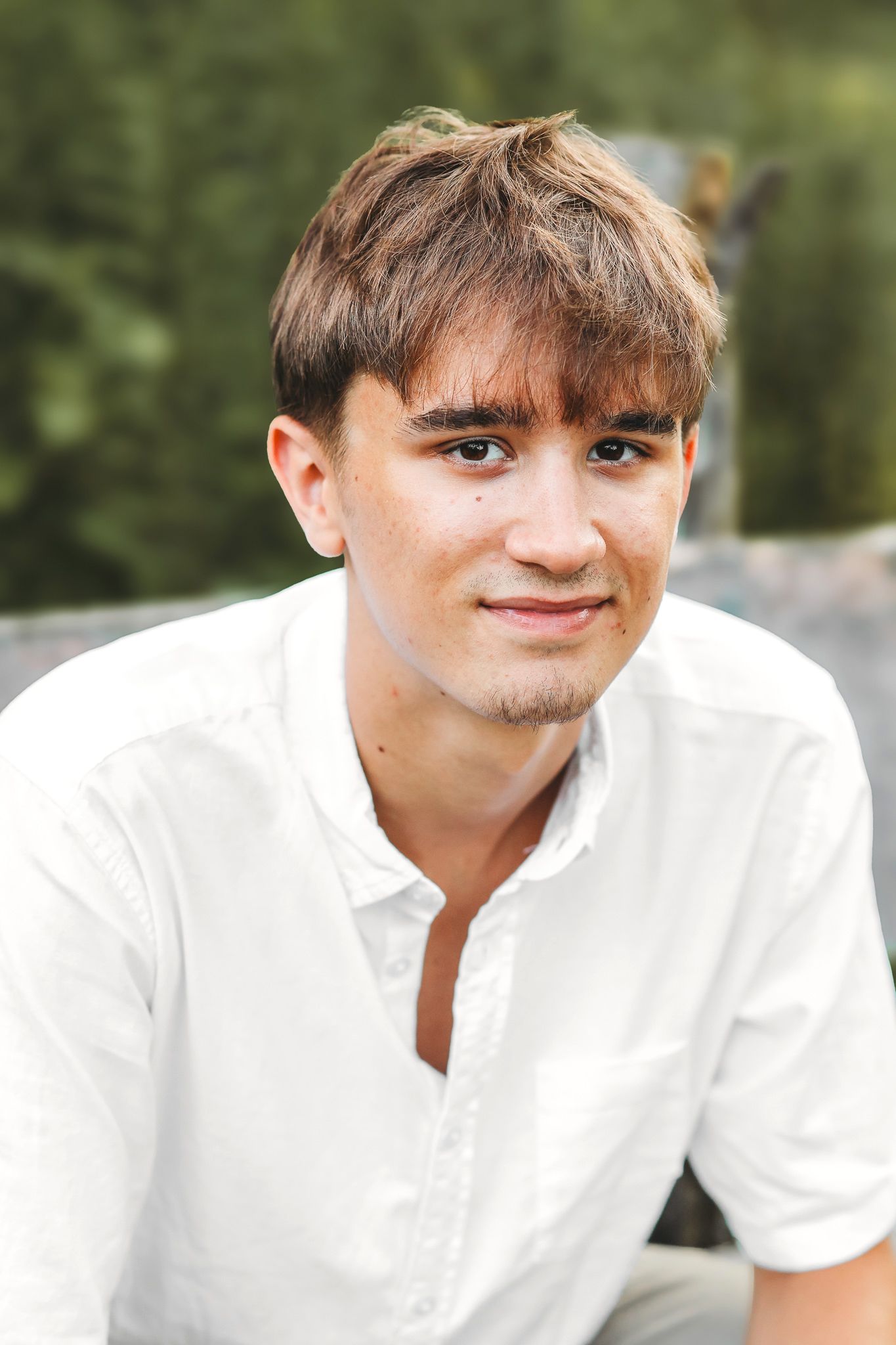 Close-up portrait of a smiling young man in a white shirt with a green outdoor background.