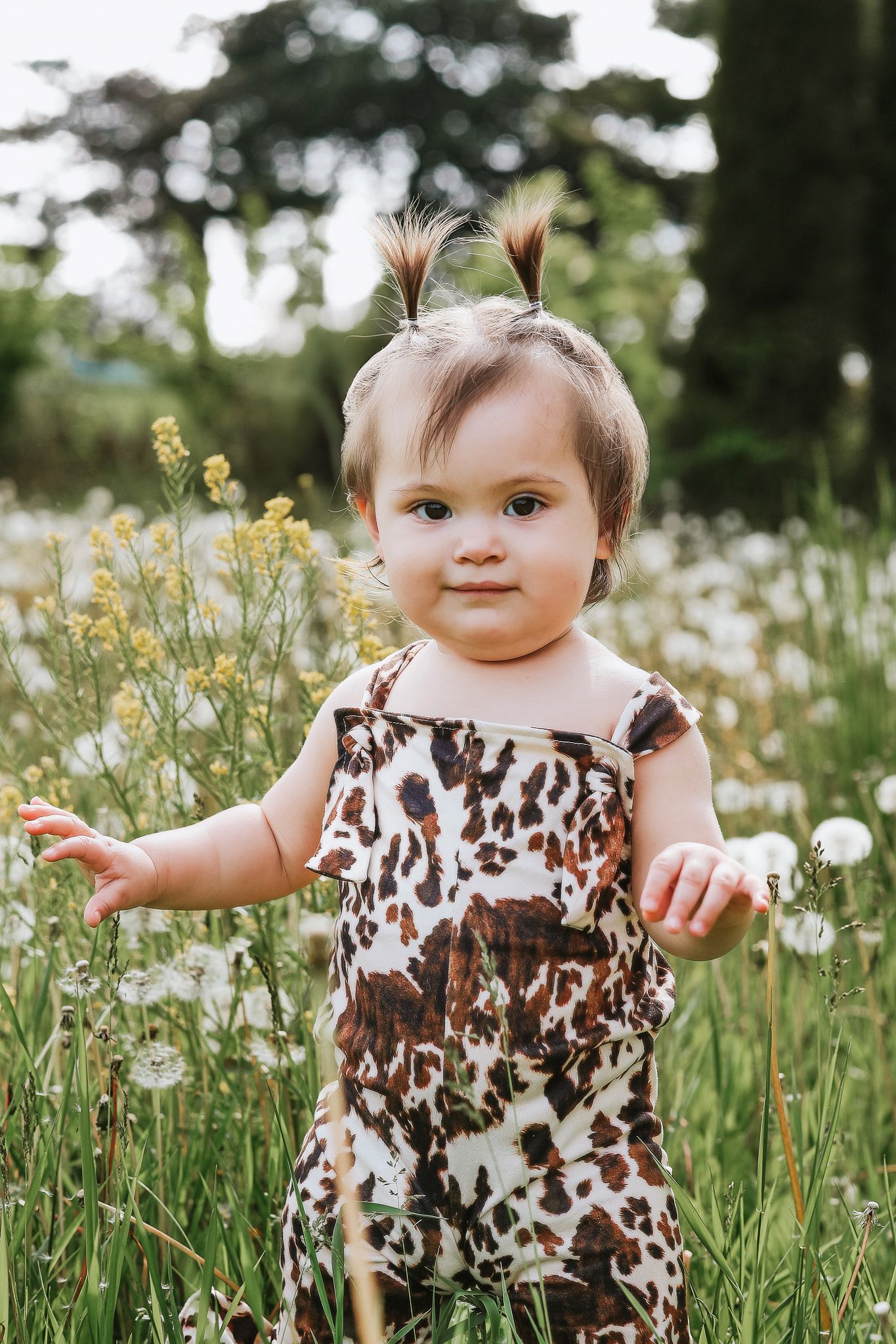 Adorable baby girl in animal print romper standing among wildflowers and dandelions outdoors.
