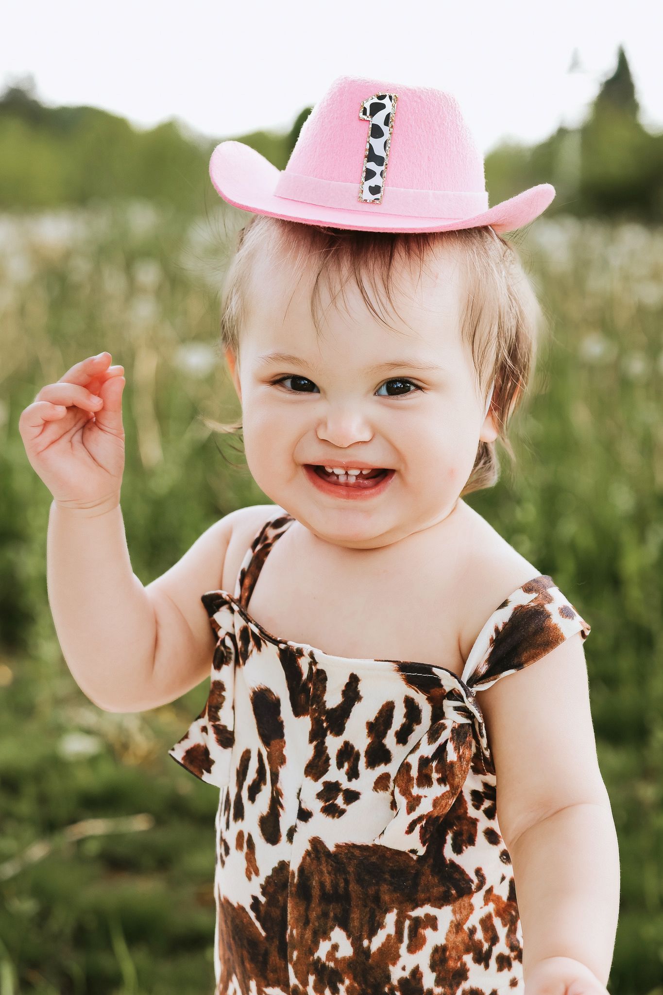 Cheerful toddler girl wearing a pink cowboy hat and cow-print dress outdoors.