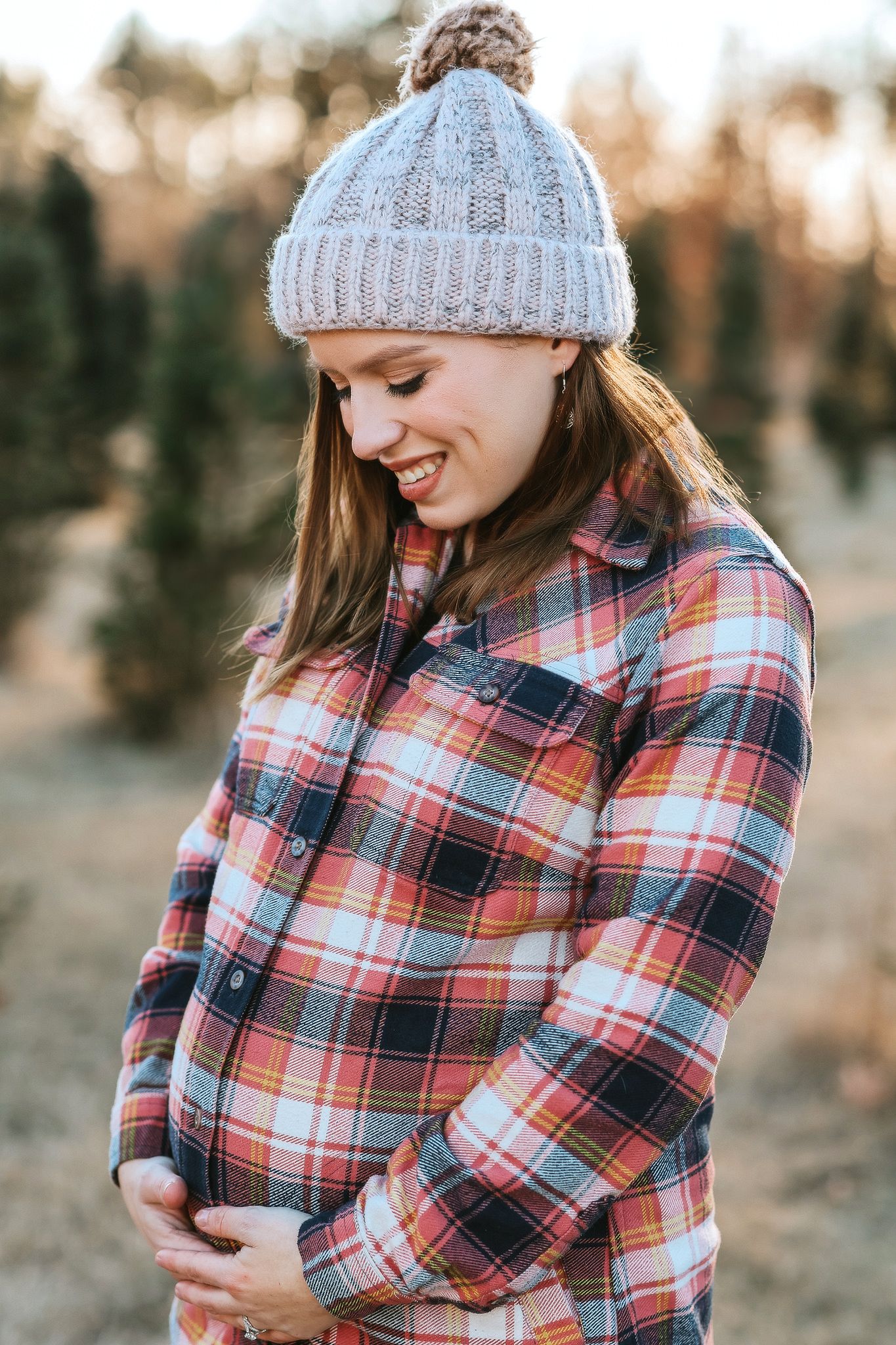 Smiling pregnant woman in plaid shirt and knit hat outdoors.