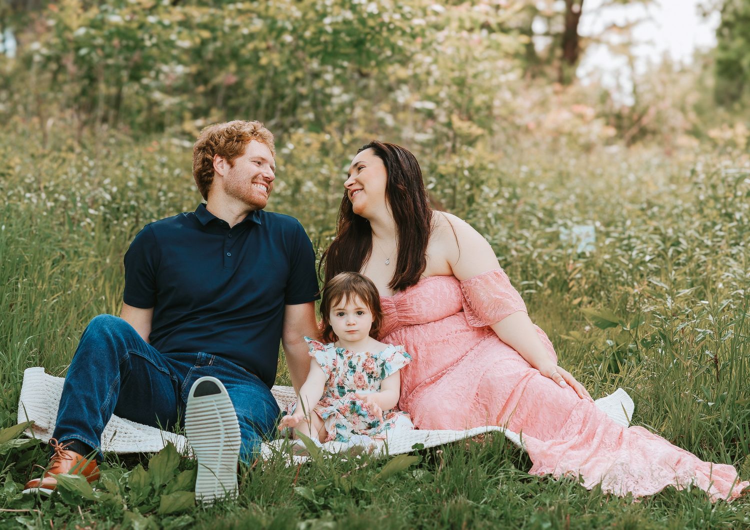 Family of three sitting in a grassy meadow, pregnant mother in pink maternity gown.