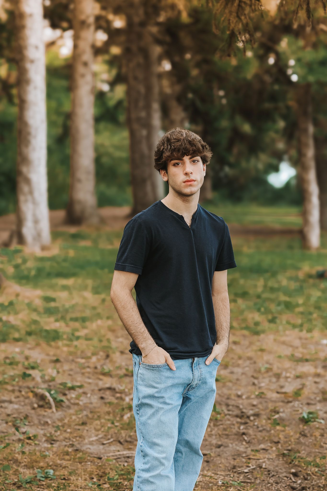 Young man in black t-shirt and jeans standing with hands in pockets among tall pine trees.
