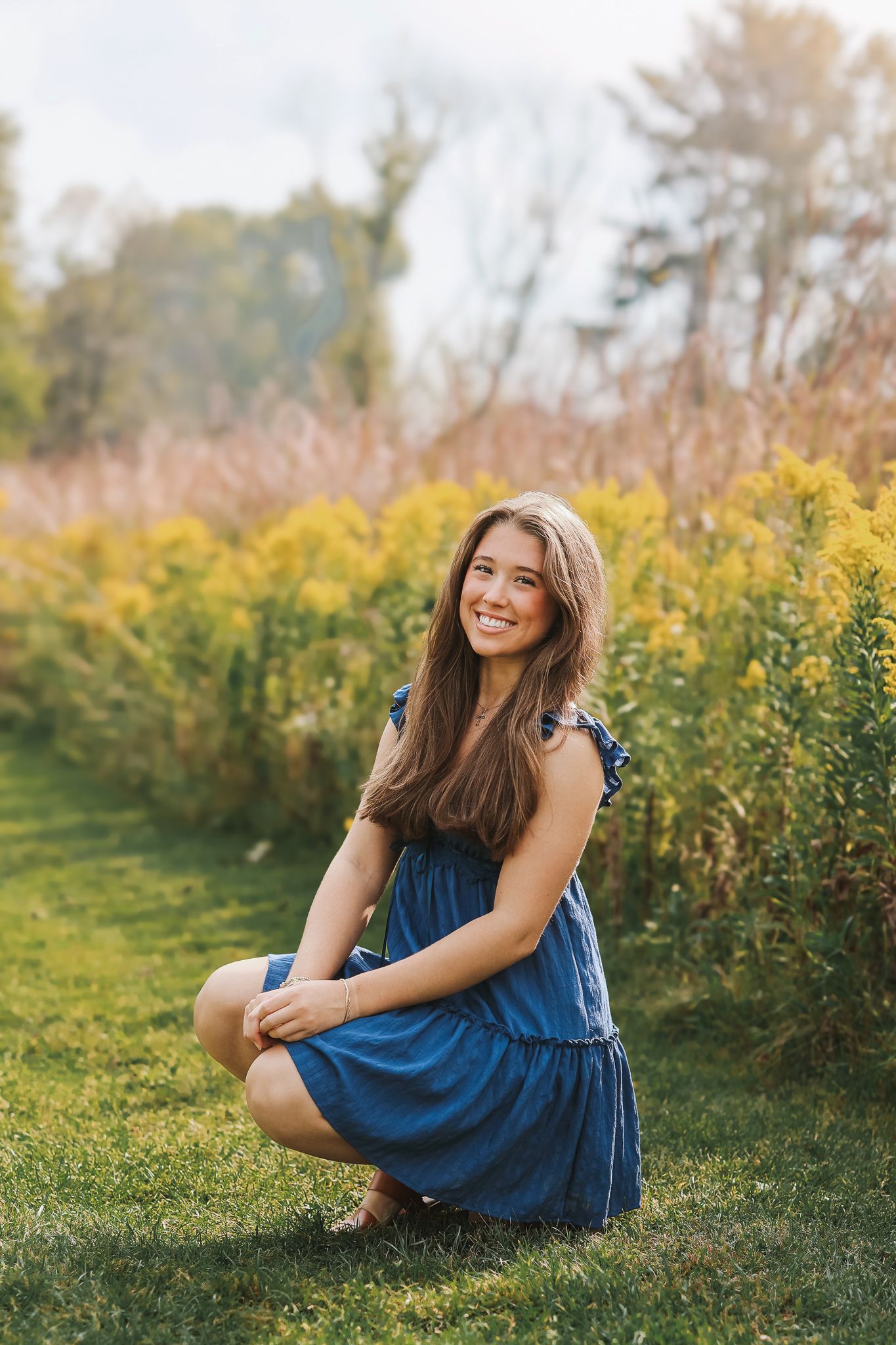 Smiling young woman in blue dress crouching on grass near yellow wildflowers outdoors.