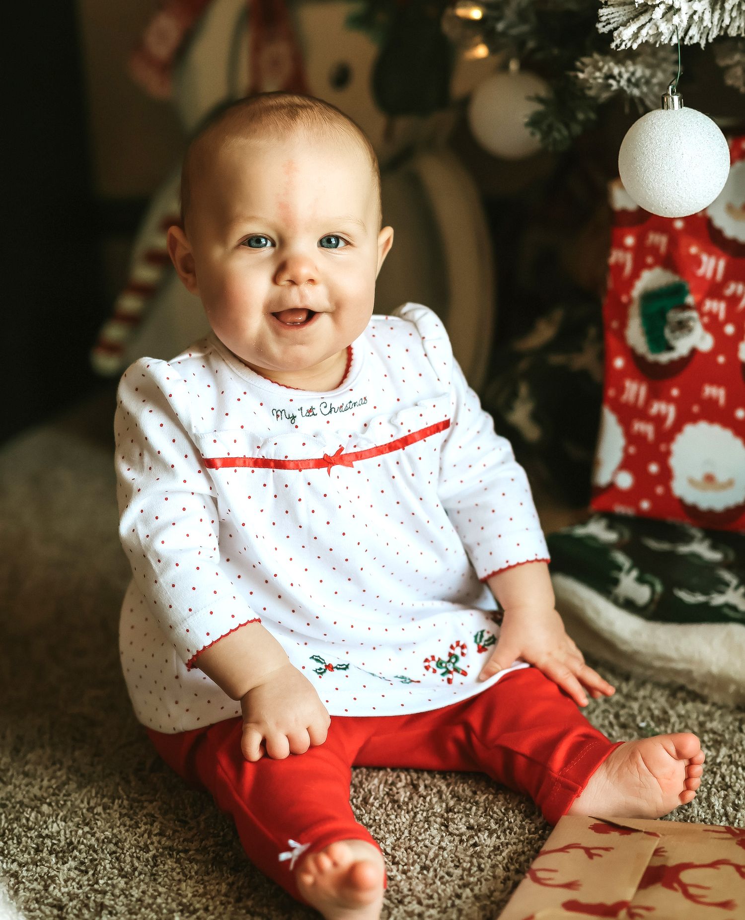 Baby girl in My 1st Christmas outfit sitting near a decorated Christmas tree smiling.