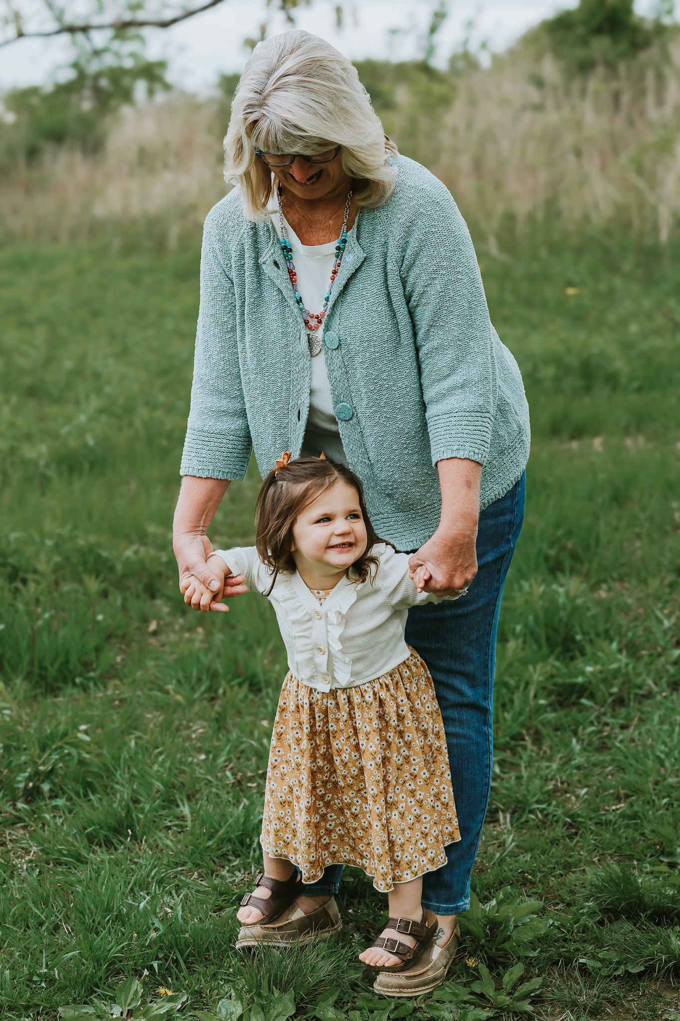 A grandmother helps a smiling toddler girl walk through a green grassy outdoor field.