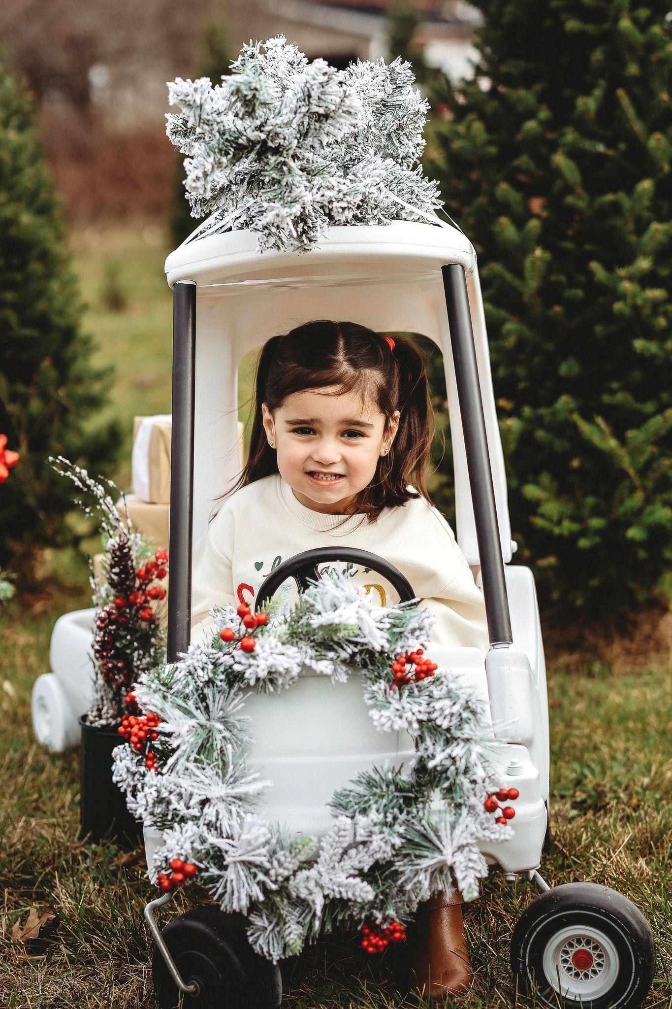 Cute little girl sitting in a white toy car decorated with a Christmas wreath and snowy tree.