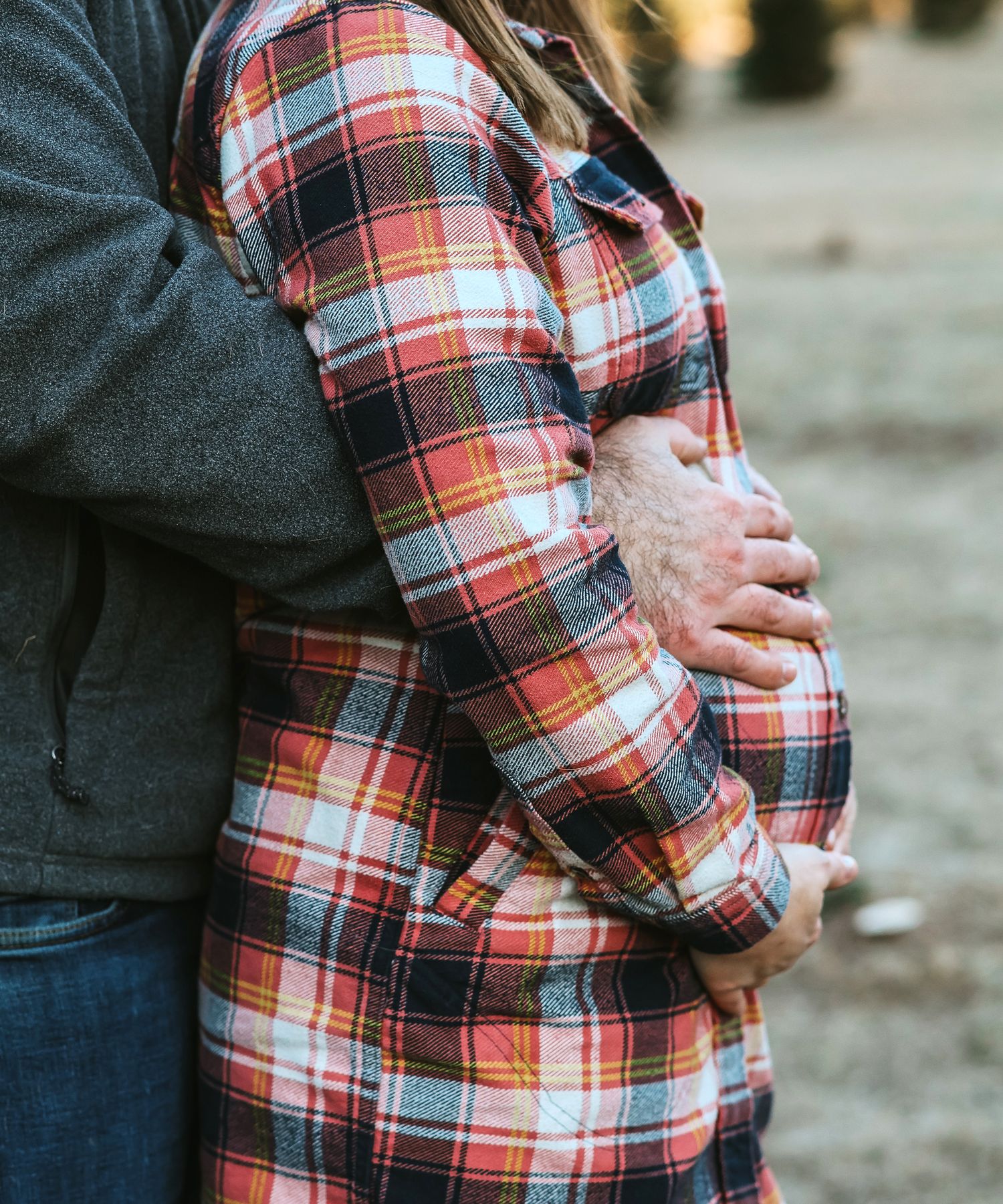 Close-up of man embracing pregnant woman in plaid shirt, hands cradling her baby bump outdoors.