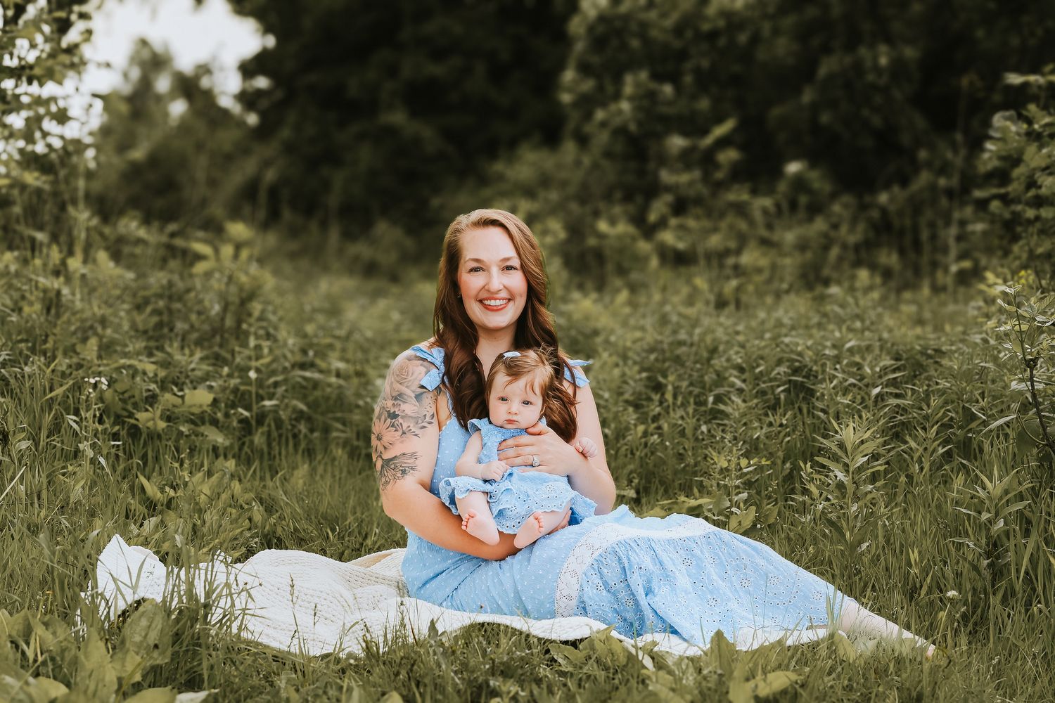 Smiling mother in blue dress holds infant baby while sitting in a lush green meadow.