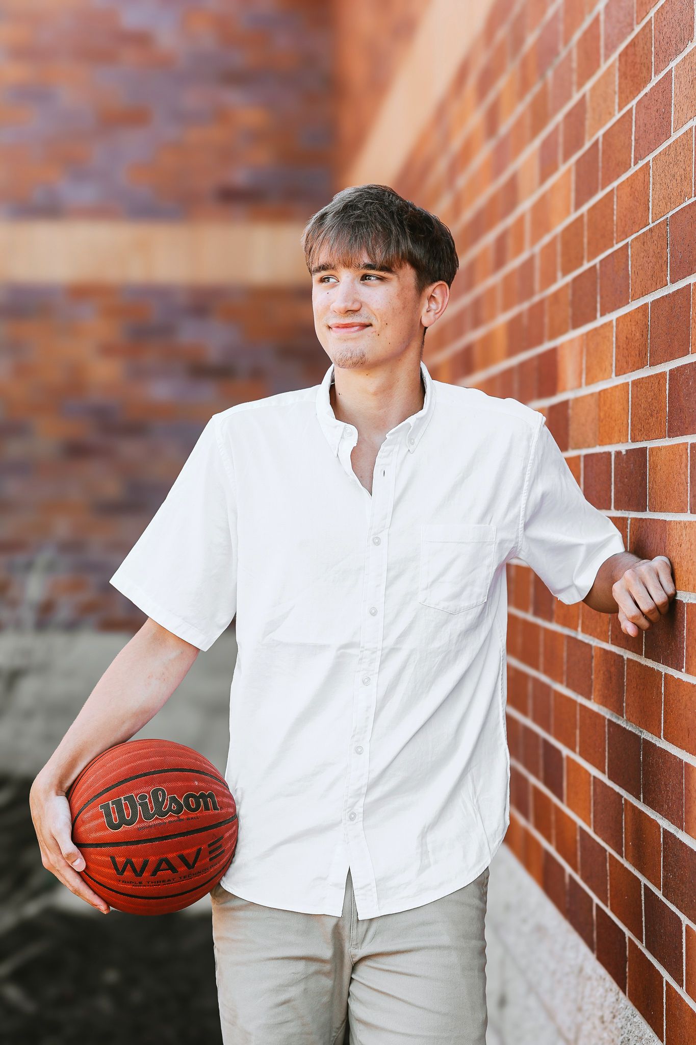 Young man in a white short-sleeve shirt holding a Wilson basketball and leaning against a red brick wall outside.