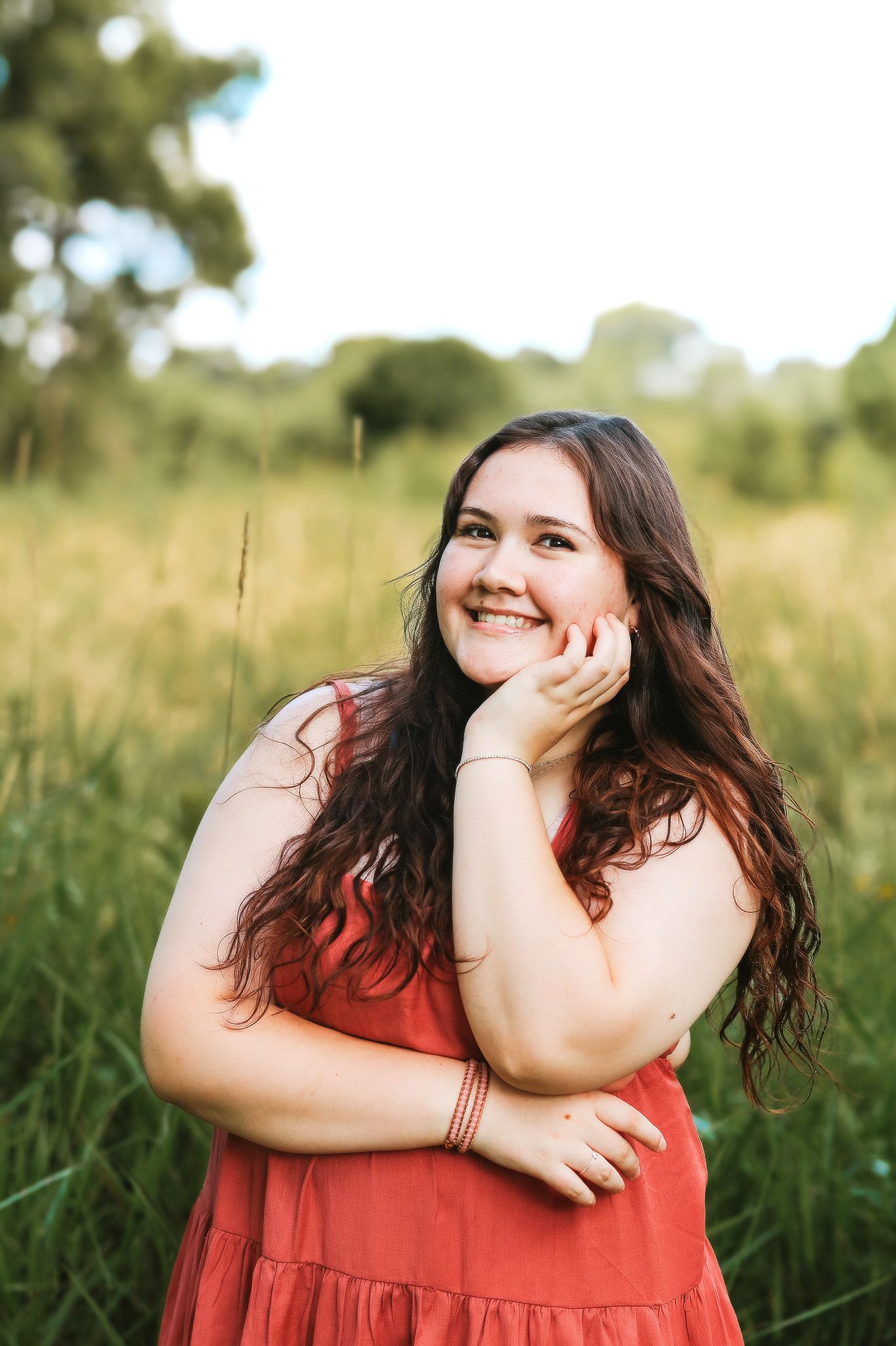 Happy young woman in red dress posing in golden grass field with hand on cheek.