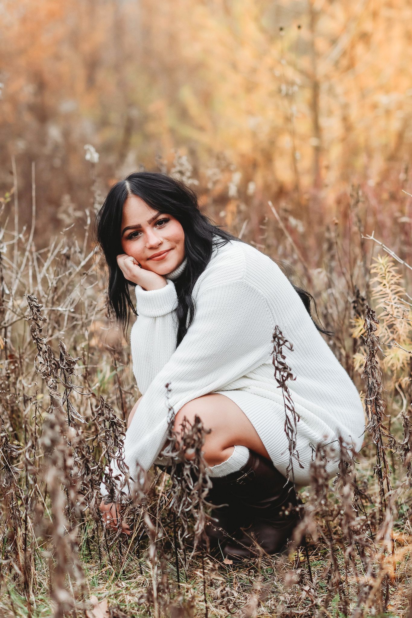Dark-haired young woman in white sweater crouching gracefully among dry autumn wildflowers in a field.
