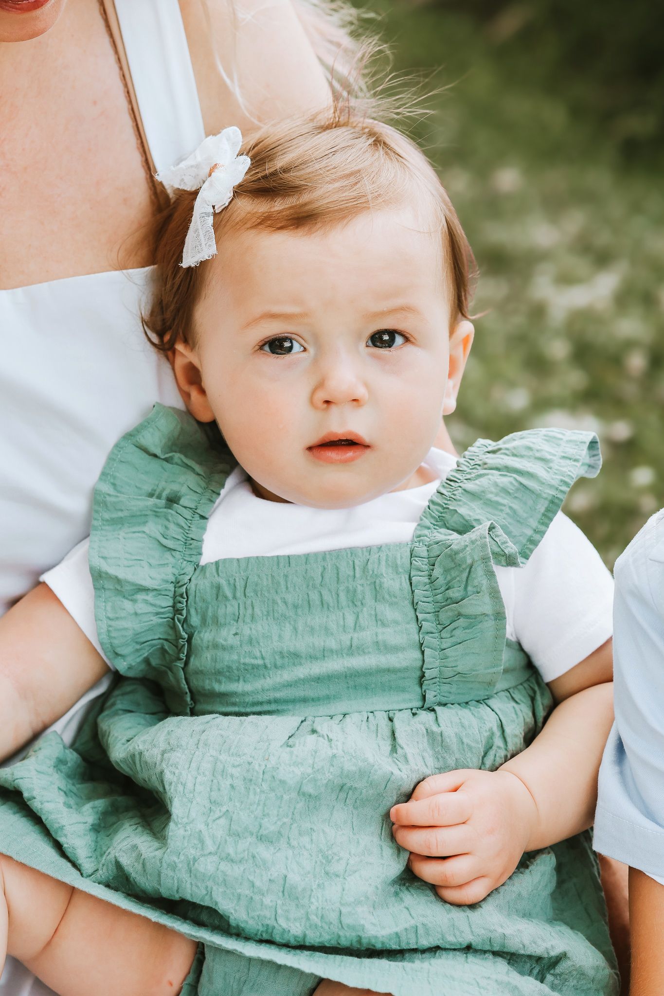 Close-up of an adorable baby girl in a green ruffle dress with a white bow, sitting outdoors.