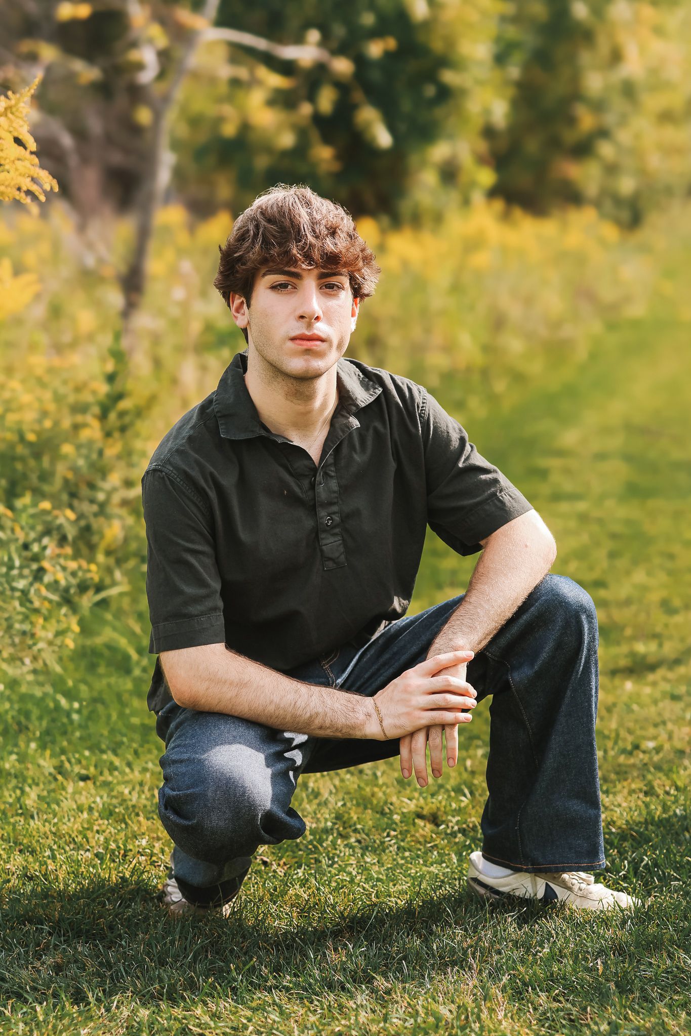 Young man in dark polo shirt and jeans crouching on grass in autumn park setting.