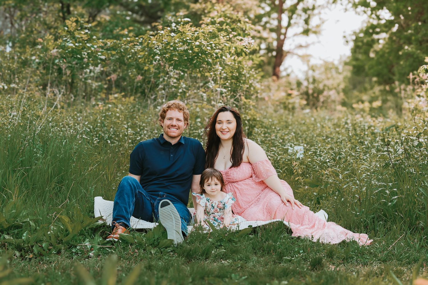 Happy family of three sitting in a wildflower meadow, mom in pink dress, dad in navy shirt, toddler between them.