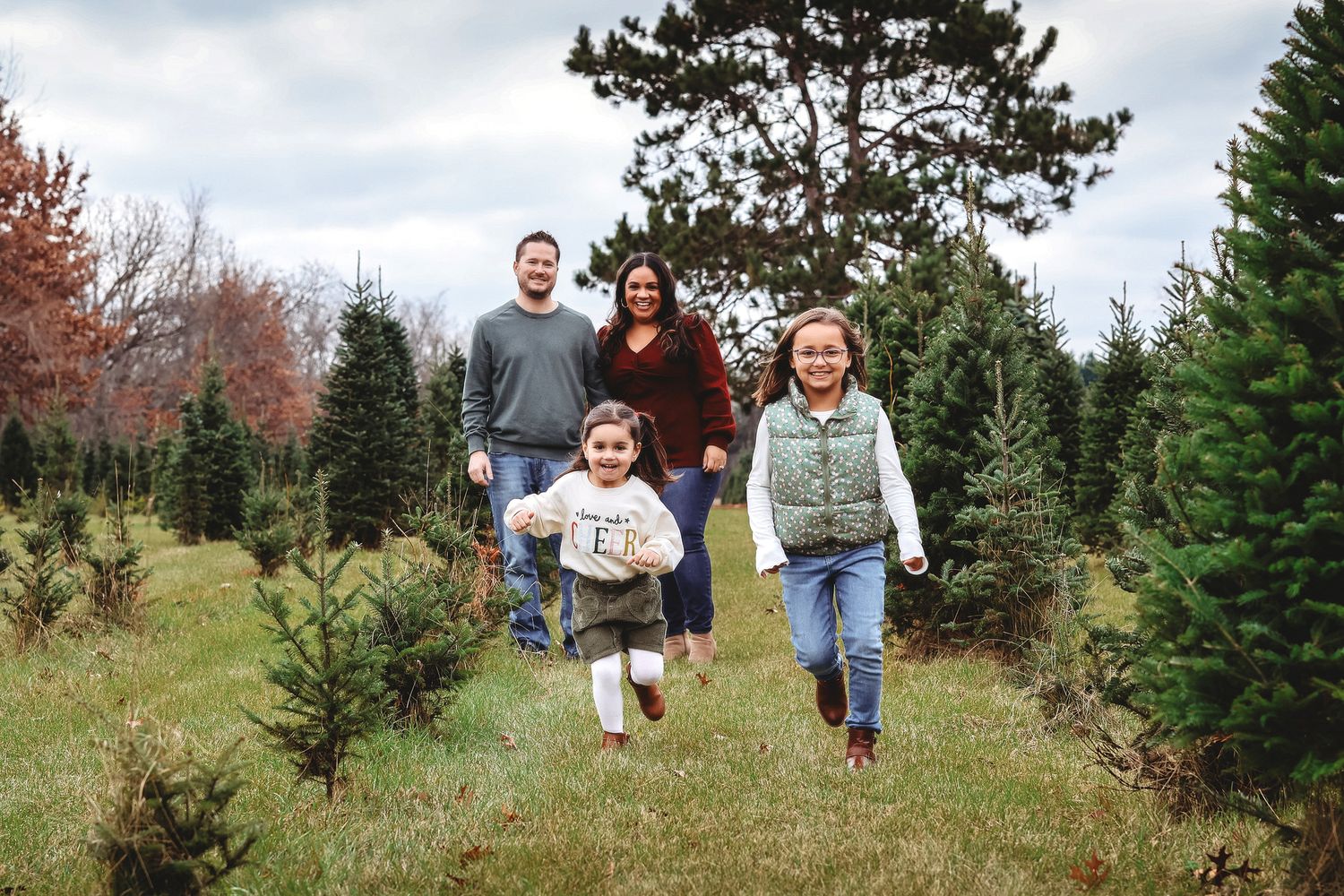 Happy family with two children running through a Christmas tree farm outdoors.