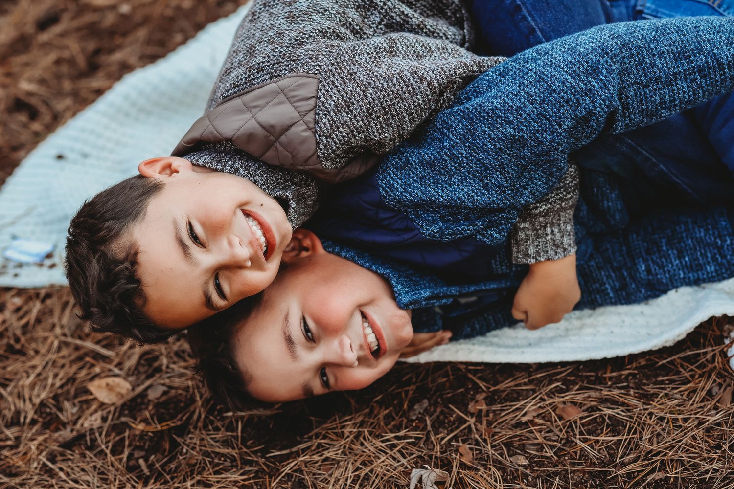 Two young brothers lying on a blanket outdoors, smiling and laughing together.