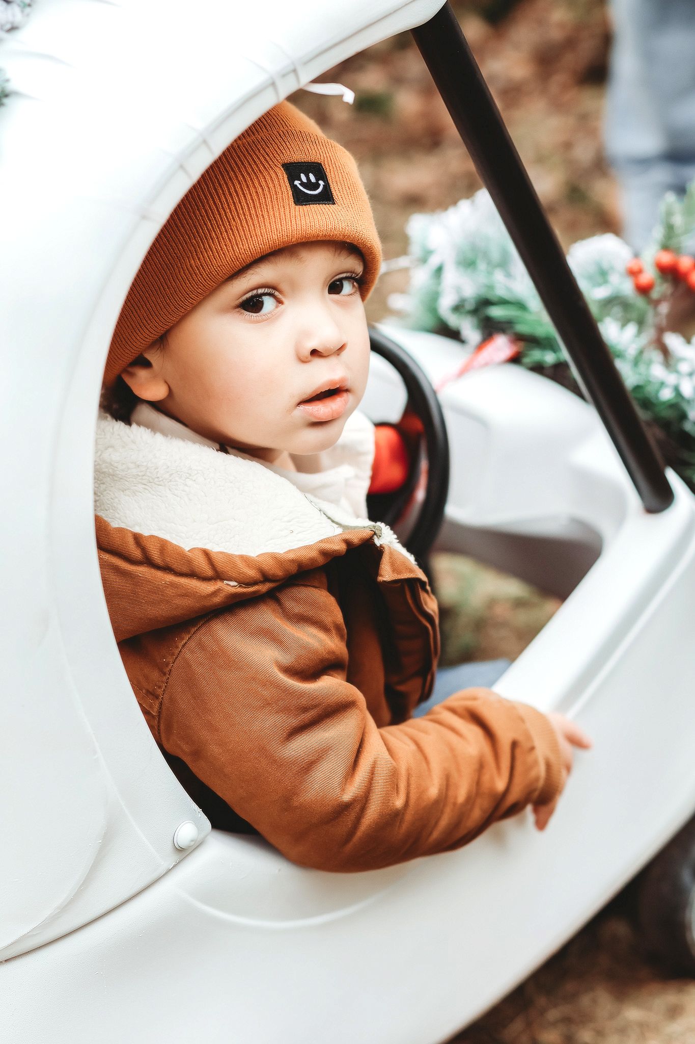 Adorable toddler in brown jacket and beanie sitting in a white toy car outdoors.