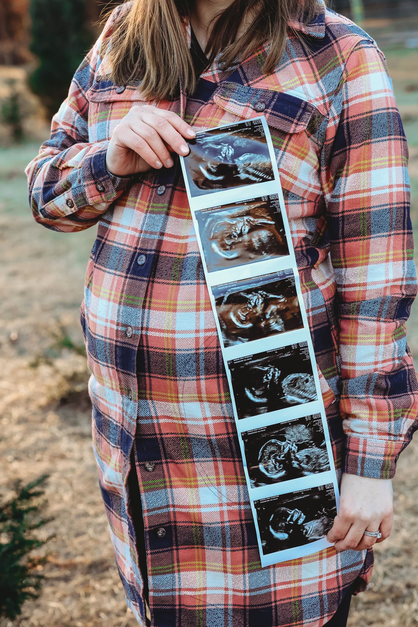 Pregnant woman in plaid shirt holding ultrasound strip photos against her baby bump outdoors.