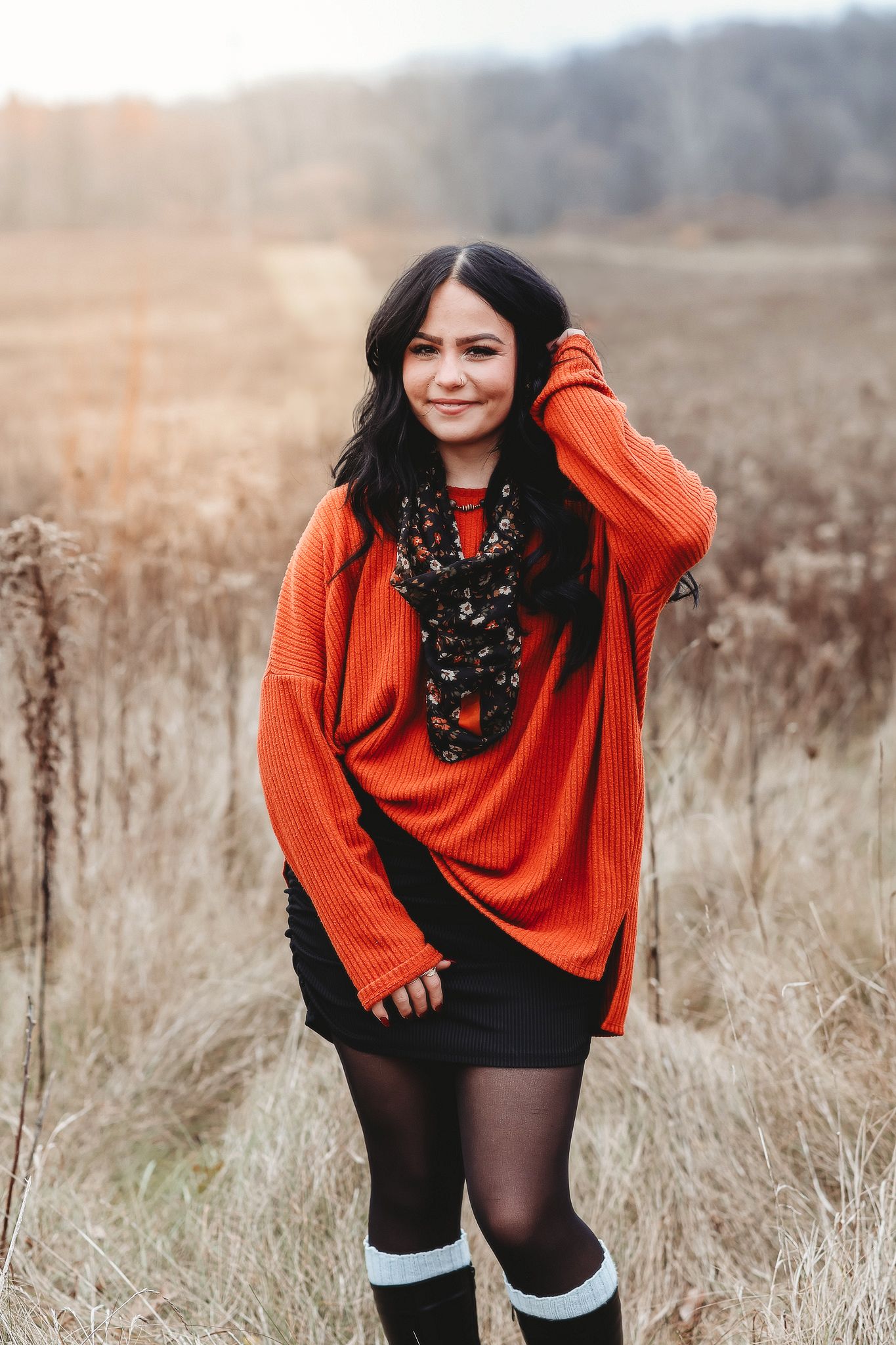 Young woman in orange sweater and black skirt standing in an autumn field with tall dry grass.