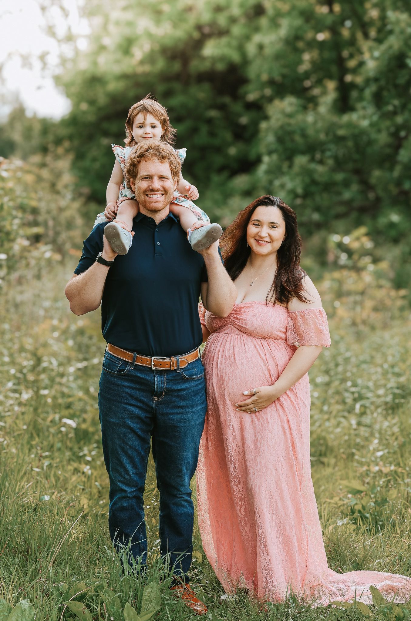 Smiling family with toddler on dads shoulders and pregnant mom in pink lace gown outdoors.