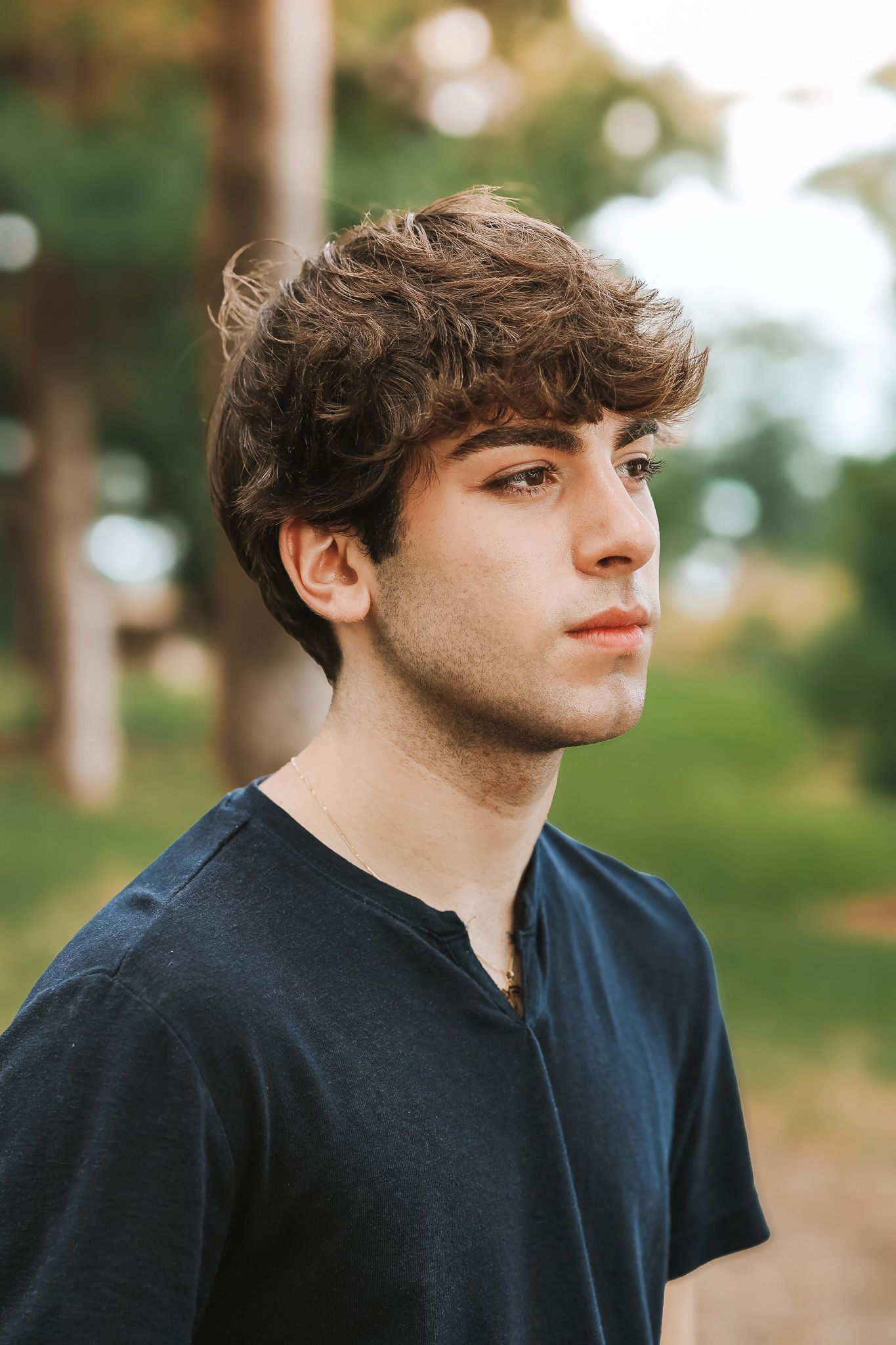 Close-up portrait of curly-haired young man in navy t-shirt with blurred trees in background.