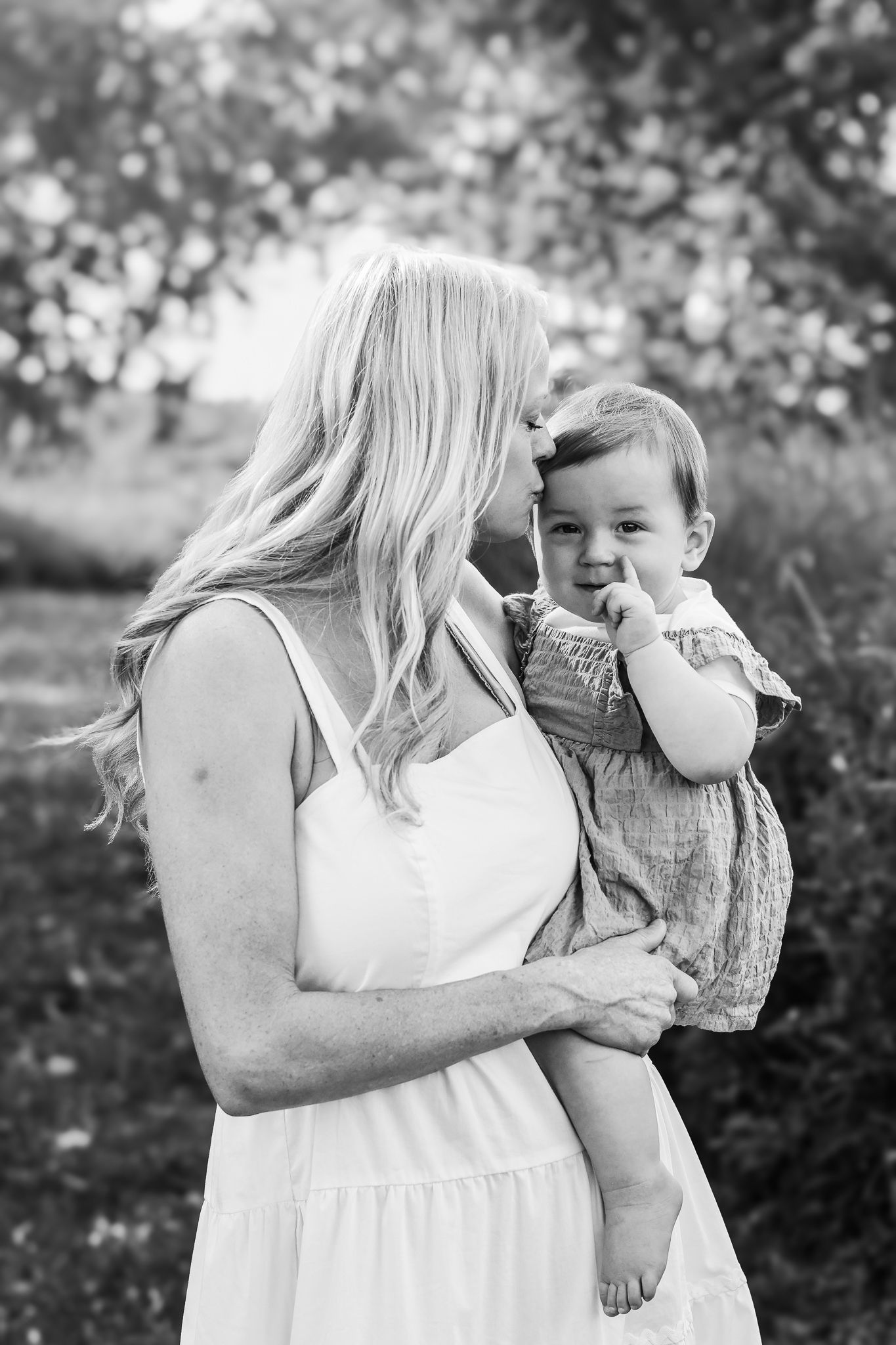 Black and white photo of a blonde mother in a white dress tenderly holding her baby girl outdoors.