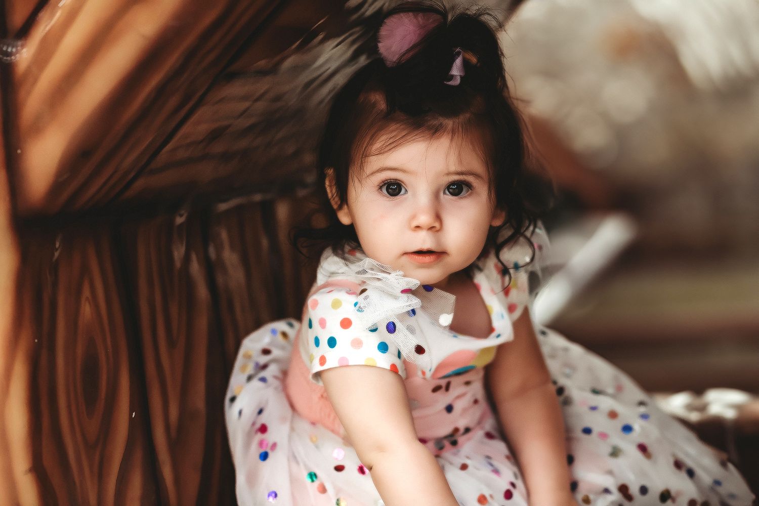 Adorable toddler girl in a polka dot dress with mouse ear hair clips sitting in a wooden structure.