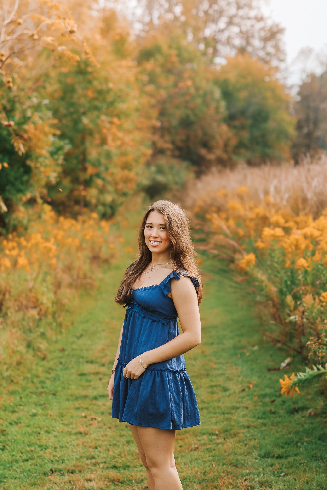 Young woman in blue dress standing on green path surrounded by golden autumn foliage.