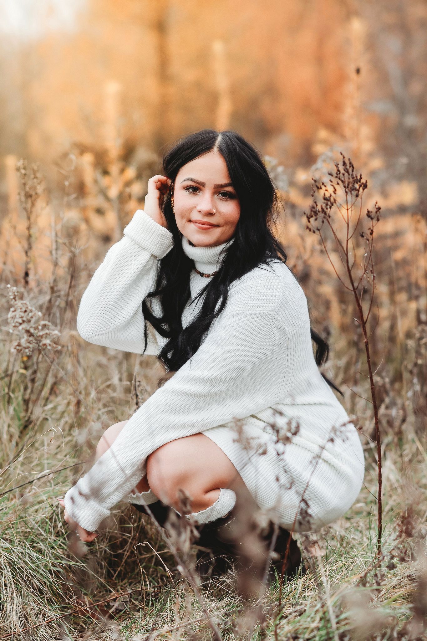Young woman with dark hair wearing a white turtleneck sweater dress posing in autumn field.
