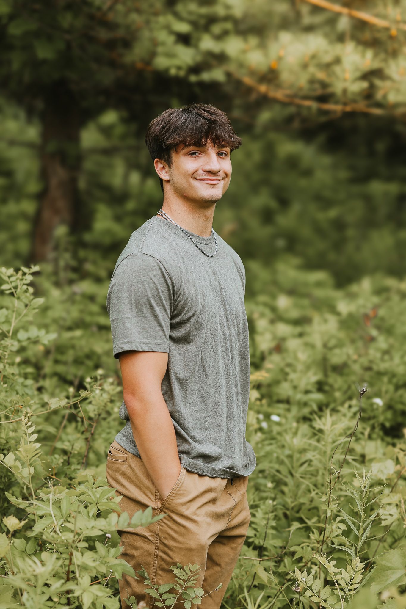 Smiling young man in gray t-shirt standing among lush green plants in nature.