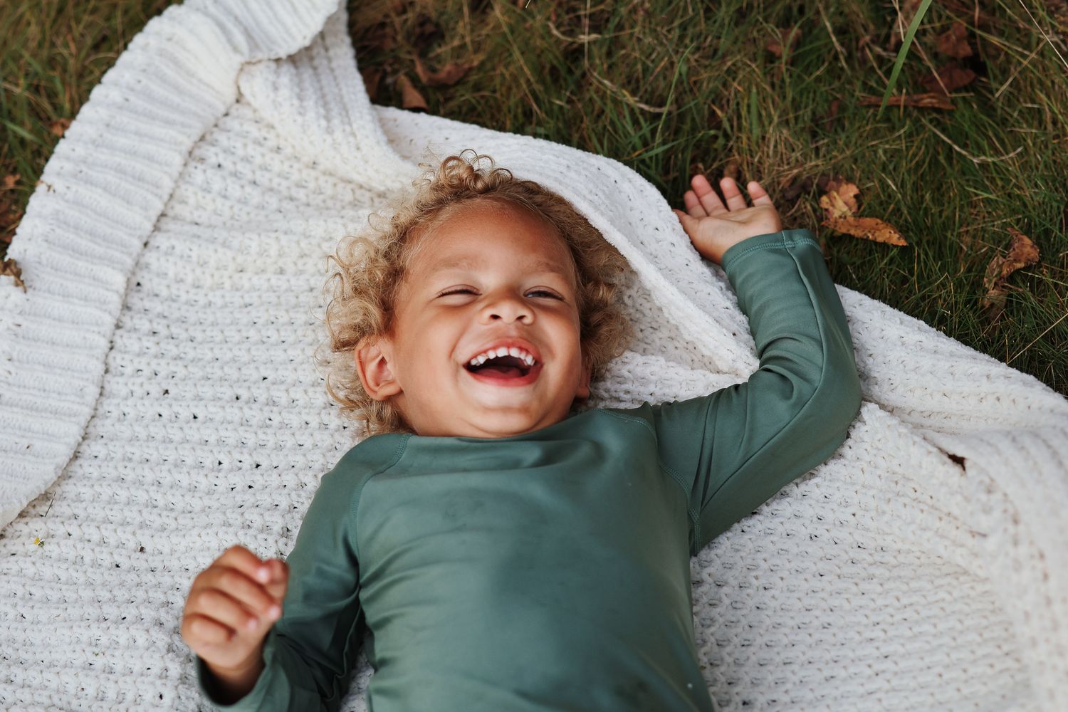 Joyful toddler with curly hair laughing while lying on a white knit blanket outdoors in autumn.