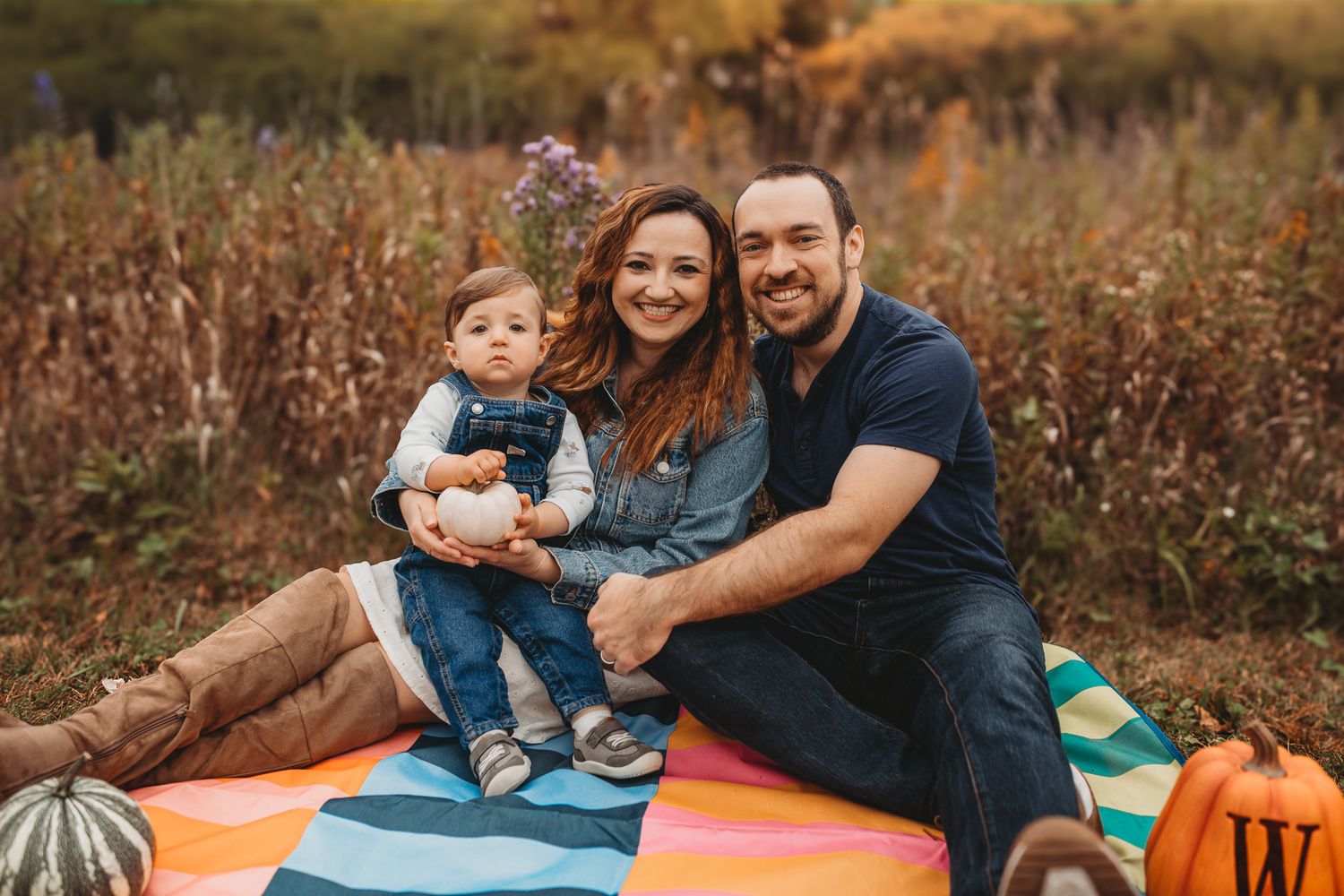 Smiling family of three poses on a colorful blanket surrounded by fall foliage and pumpkins.